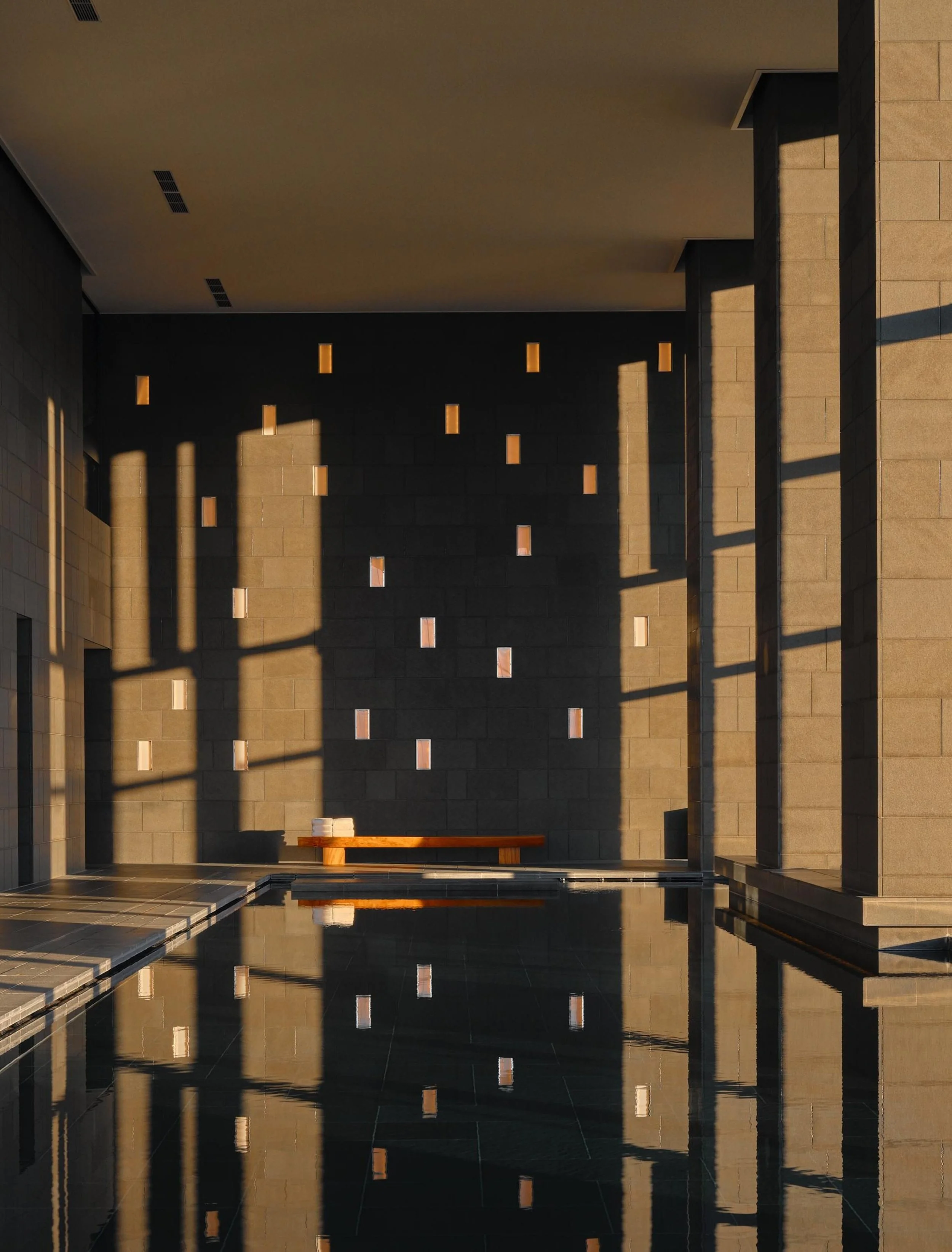 An indoor space with beige walls featuring rectangular cutouts, a reflective pool or floor, and vertical beams casting shadows, with a wooden bench and folded towels.