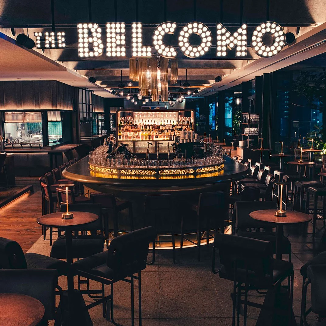A dimly lit bar with a large illuminated sign saying 'THE BELCOMO' hanging from the ceiling. The bar features a curved counter lined with glasses and behind it, shelves stocked with bottles. The seating area has small round tables with chairs, and there are decorative lights and plants around the space.