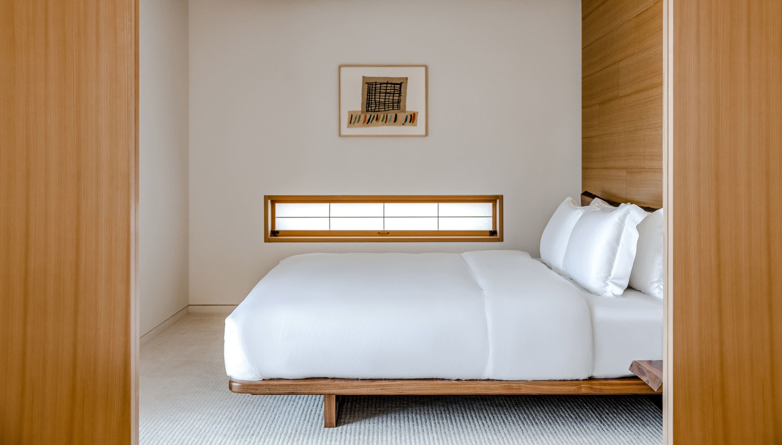 A minimalistic bedroom with a white bed and white pillows, wooden accents on the bed frame and headboard, a narrow horizontal window behind the bed, and a framed abstract artwork on the wall.