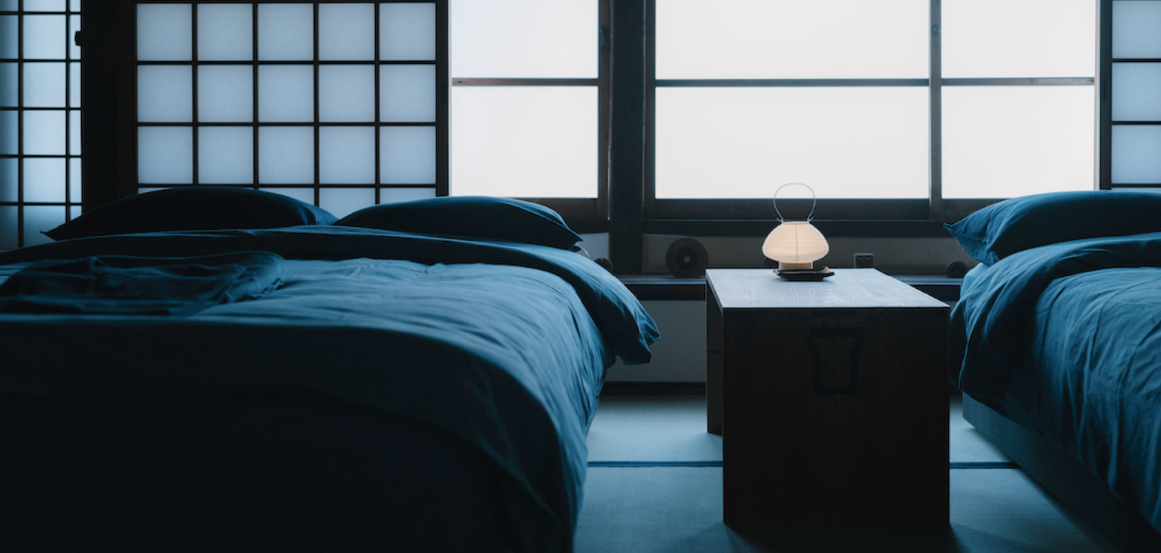 Two beds with dark bedding and pillows, a nightstand with a lamp between them, large window with traditional Japanese shoji screens in the background.