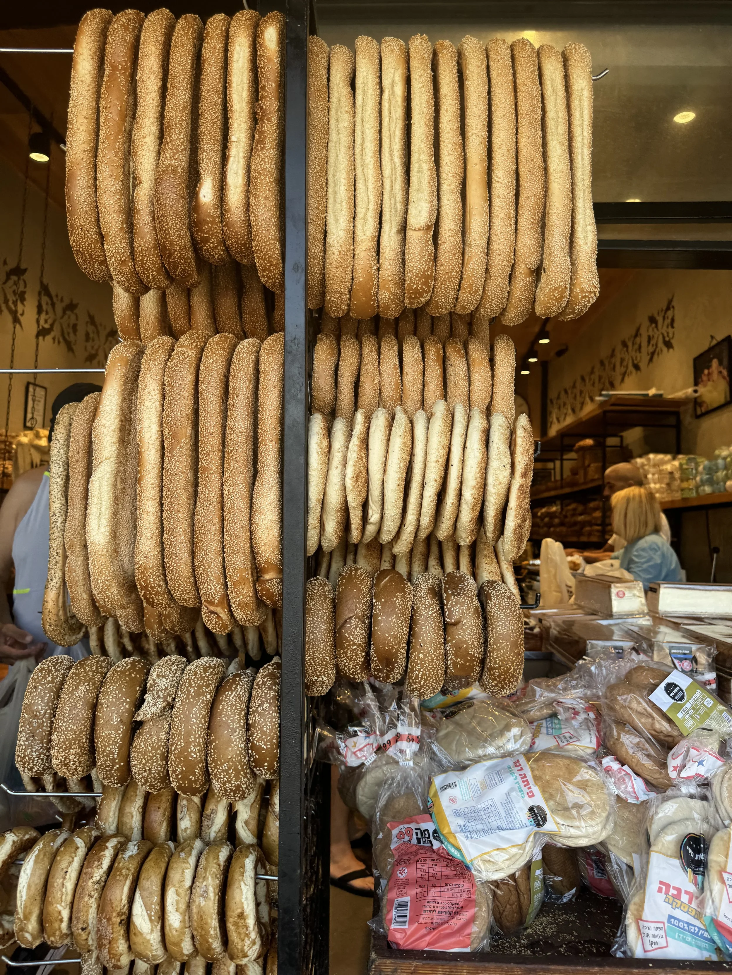 Display of various types of sesame seed bagels hanging on a metal rack in a bakery.