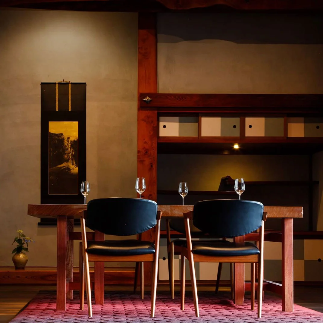 A dining table with four wine glasses in a cozy, dimly lit room featuring wooden furniture and a decorative Japanese-style wall hanging.