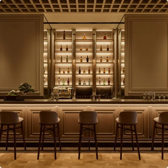 Bar counter with five chairs in front, backlit shelves with various bottles, decorative items, and glasses on the counter.