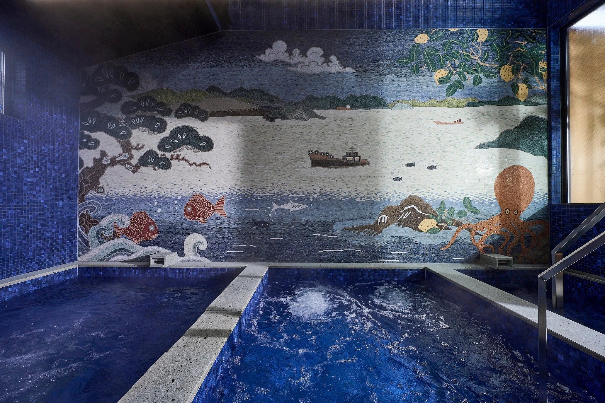 Indoor hot tub with a mosaic mural of a river scene, featuring fish, an octopus, trees, and mountains.