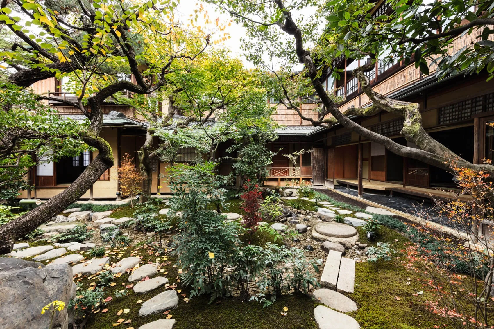 Traditional Japanese garden with winding stone paths, lush green trees, and a wooden building with sliding doors and a balcony.