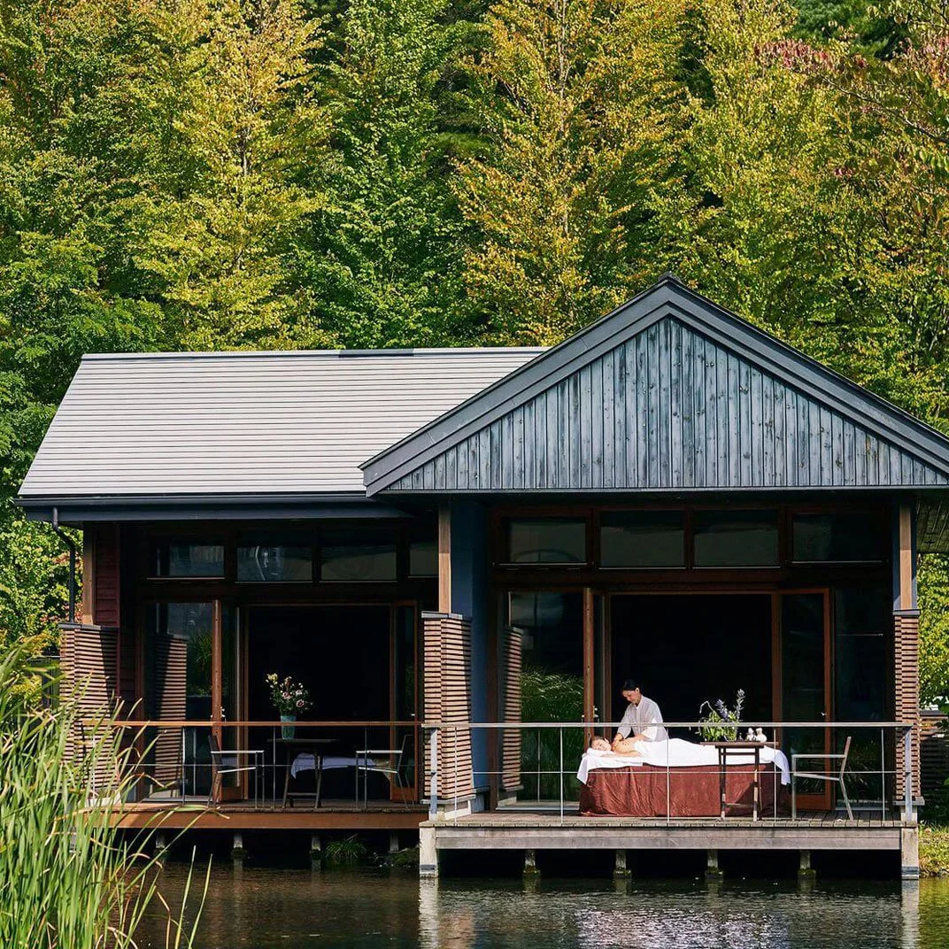 A modern house with large glass windows and a sloped gray roof, situated by a body of water, with trees in the background. On the house's deck, a person is giving a massage to another person lying on a massage table.