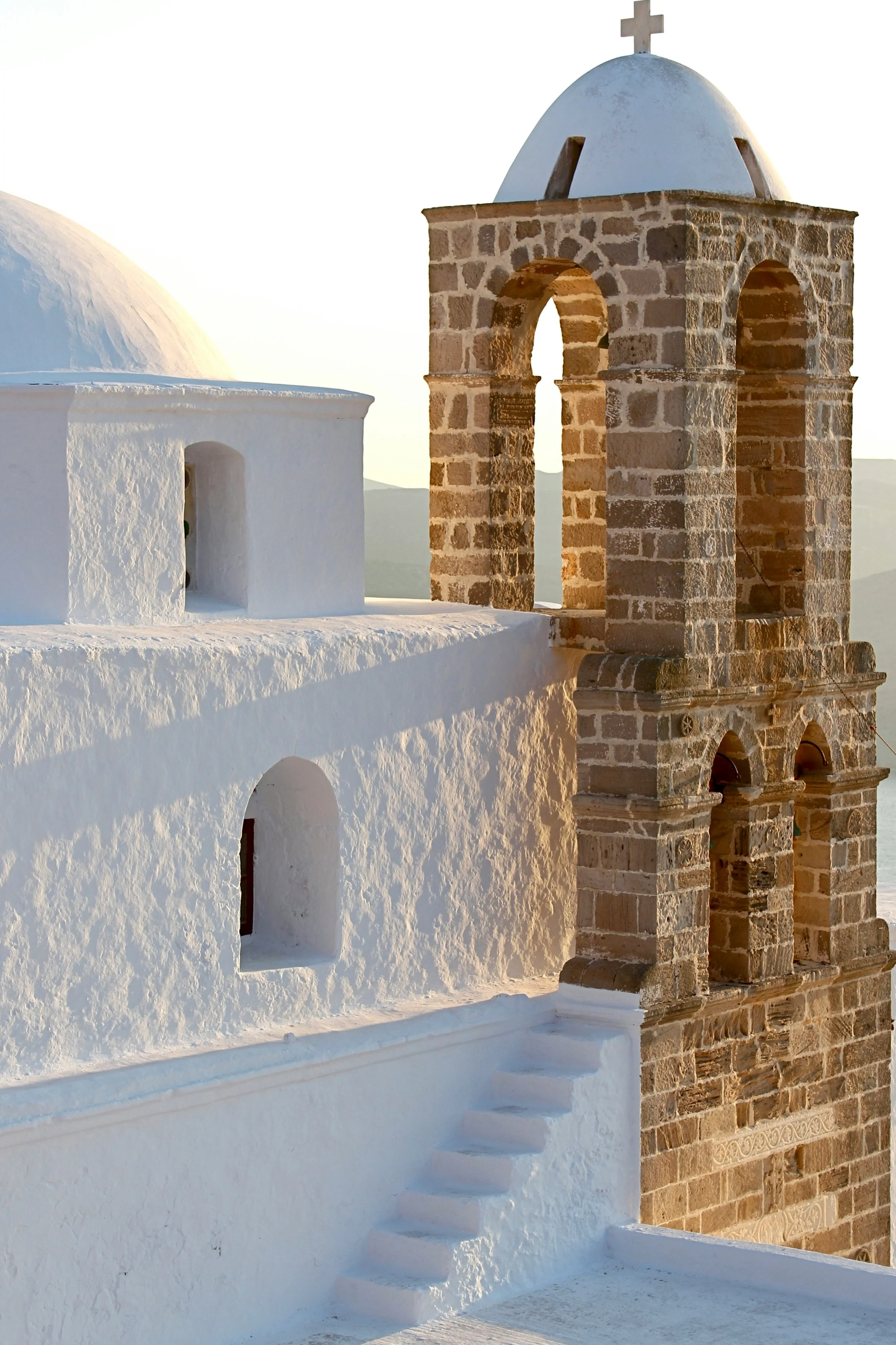 Whitewashed building with arched windows and a stone bell tower topped with a cross, typical of Greek island architecture.
