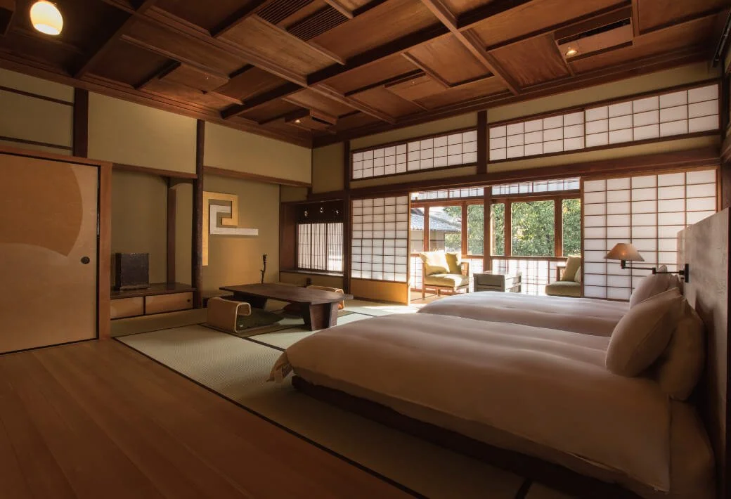 Traditional Japanese bedroom with tatami mats, sliding shoji doors, two beds, and a sitting area with chairs and a table near large windows.