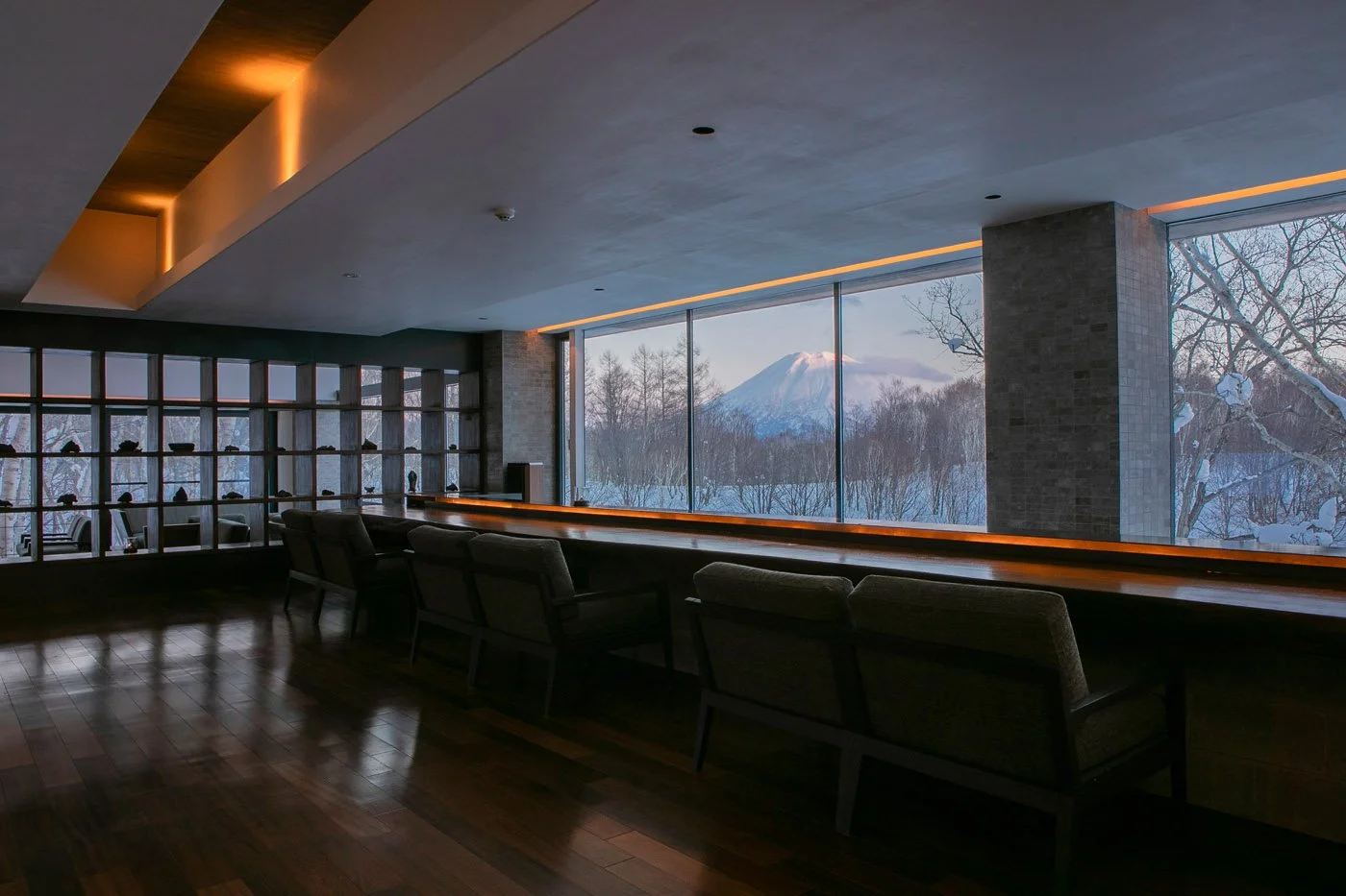 Interior of a modern lounge with a long window view of snow-covered trees and a mountain in the distance, featuring dark wooden floors, chairs, and warm lighting accents.