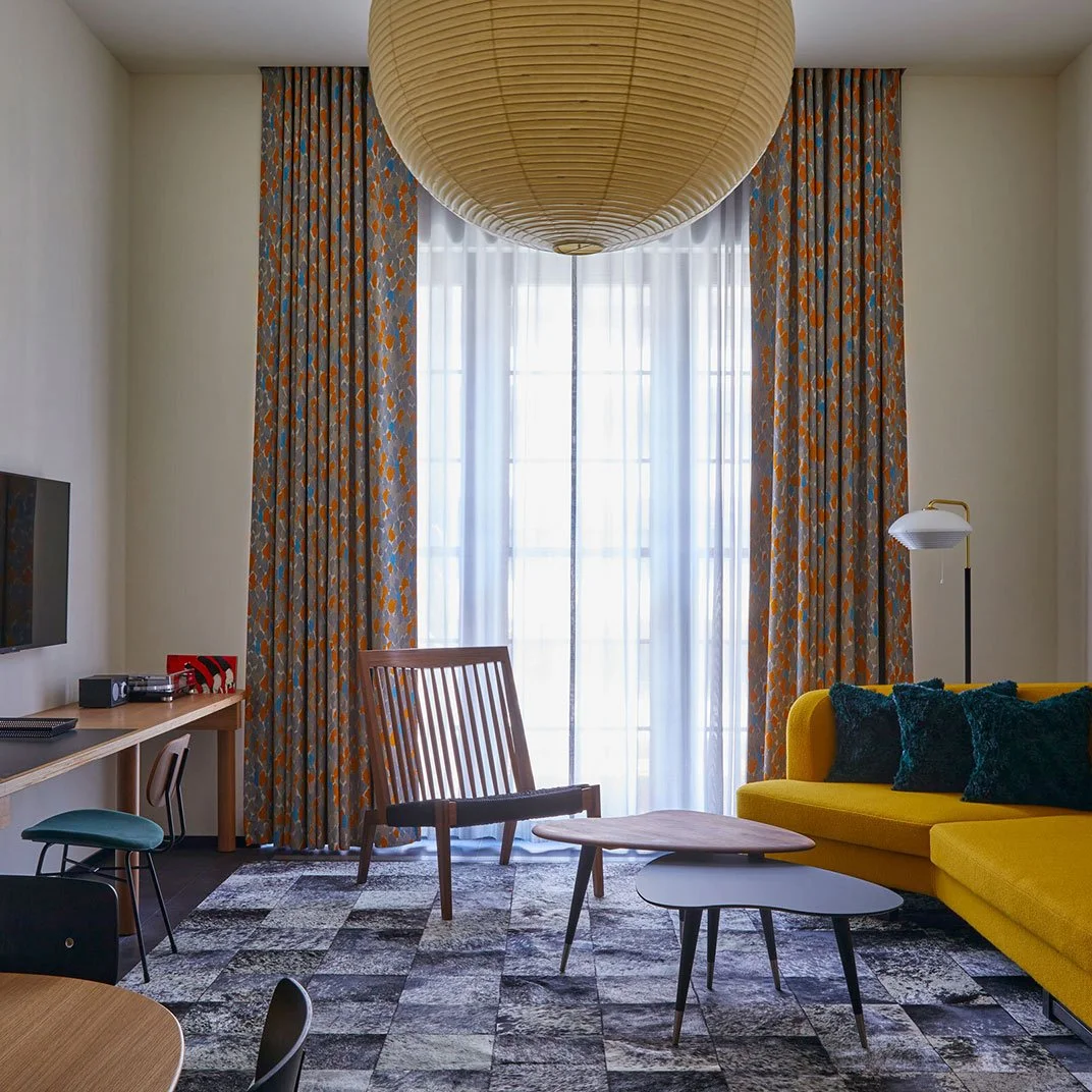 Living room with a yellow sofa, wooden armchair, two nested coffee tables, patterned curtains, and pendant ceiling light.