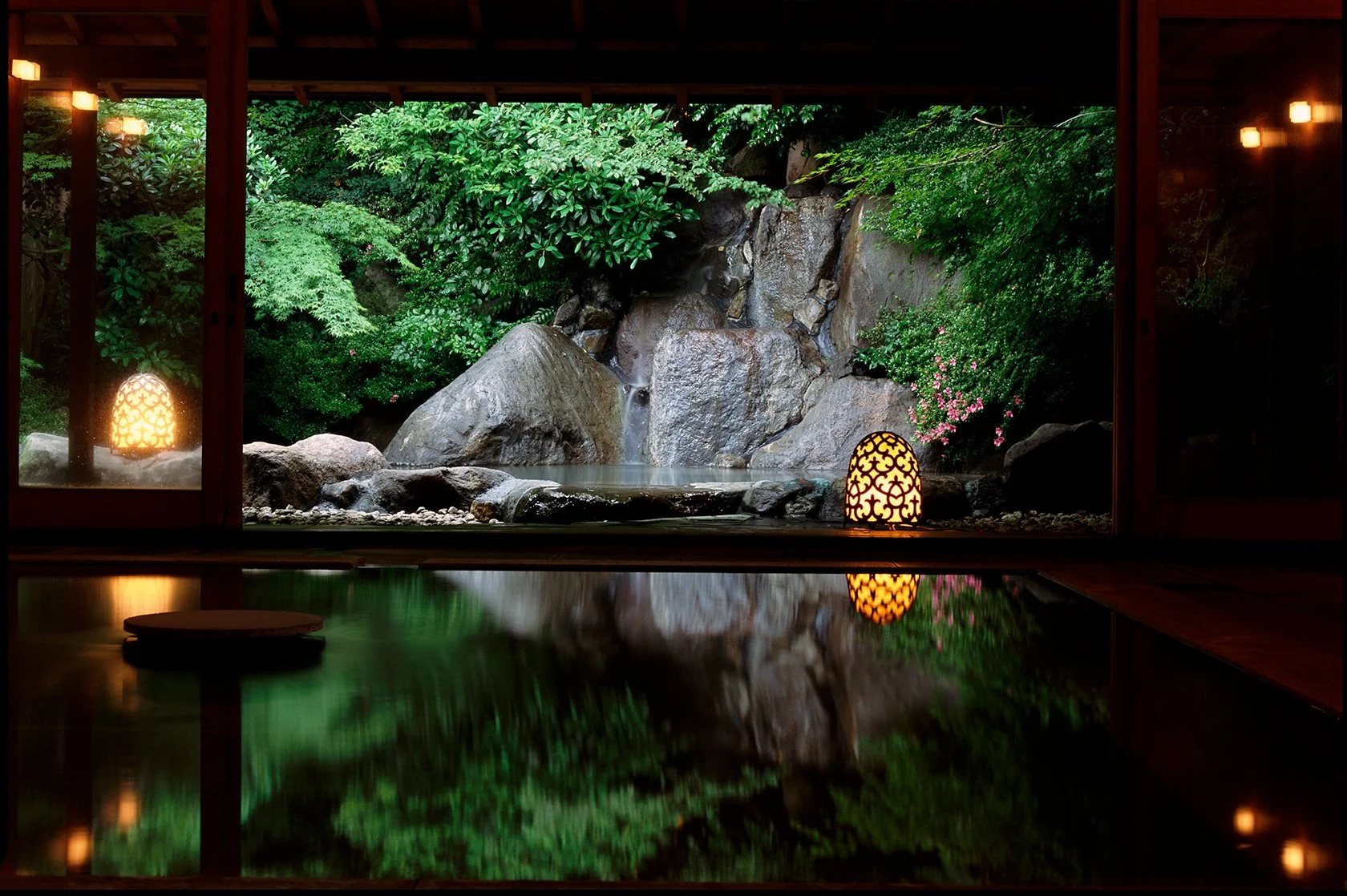Indoor view of a tranquil Japanese-style garden with large rocks, lush green plants, and small pink flowers, seen through a window at night with warm-lit decorative lanterns.