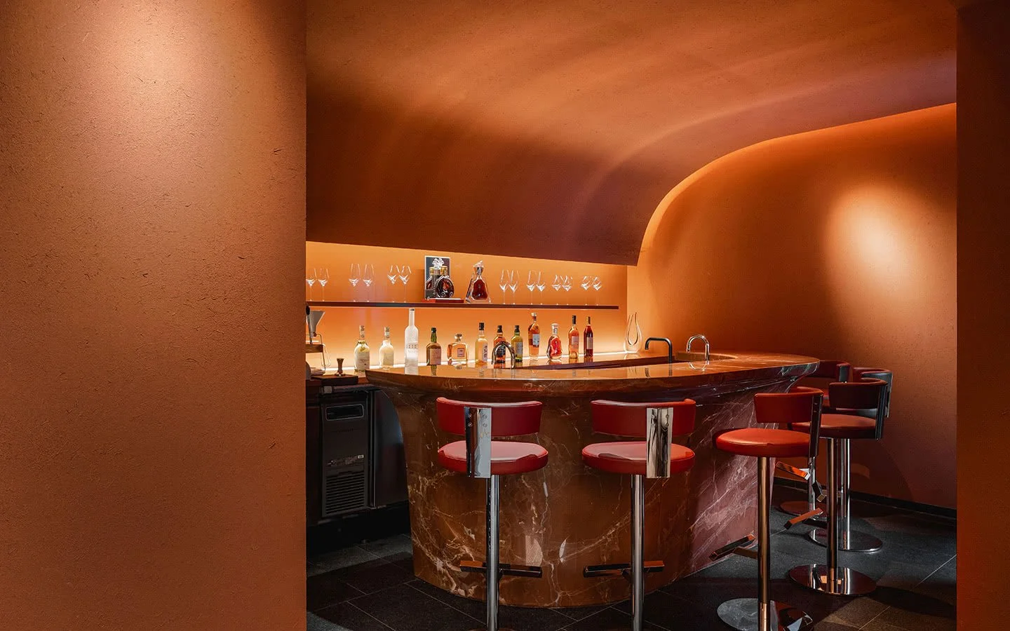 A modern bar with a curved marble counter, red bar stools, and shelves with various liquor bottles and glassware, illuminated by warm lighting.