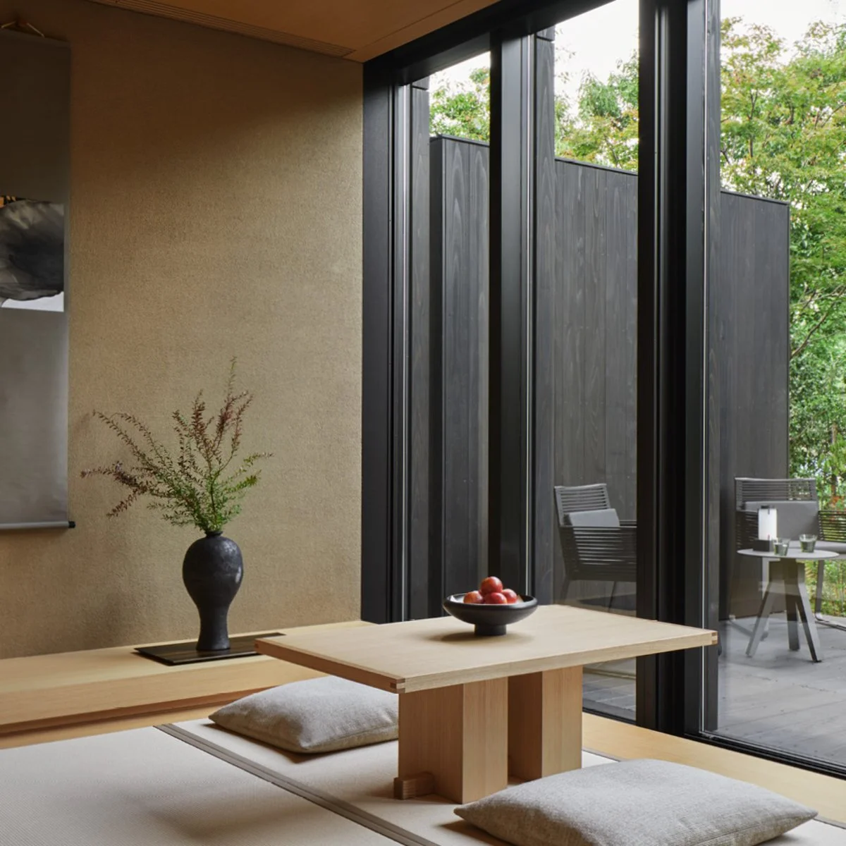 Interior of a minimalist room with a wooden table, cushions, a black vase with greenery, and a view of an outdoor balcony with chairs and trees.