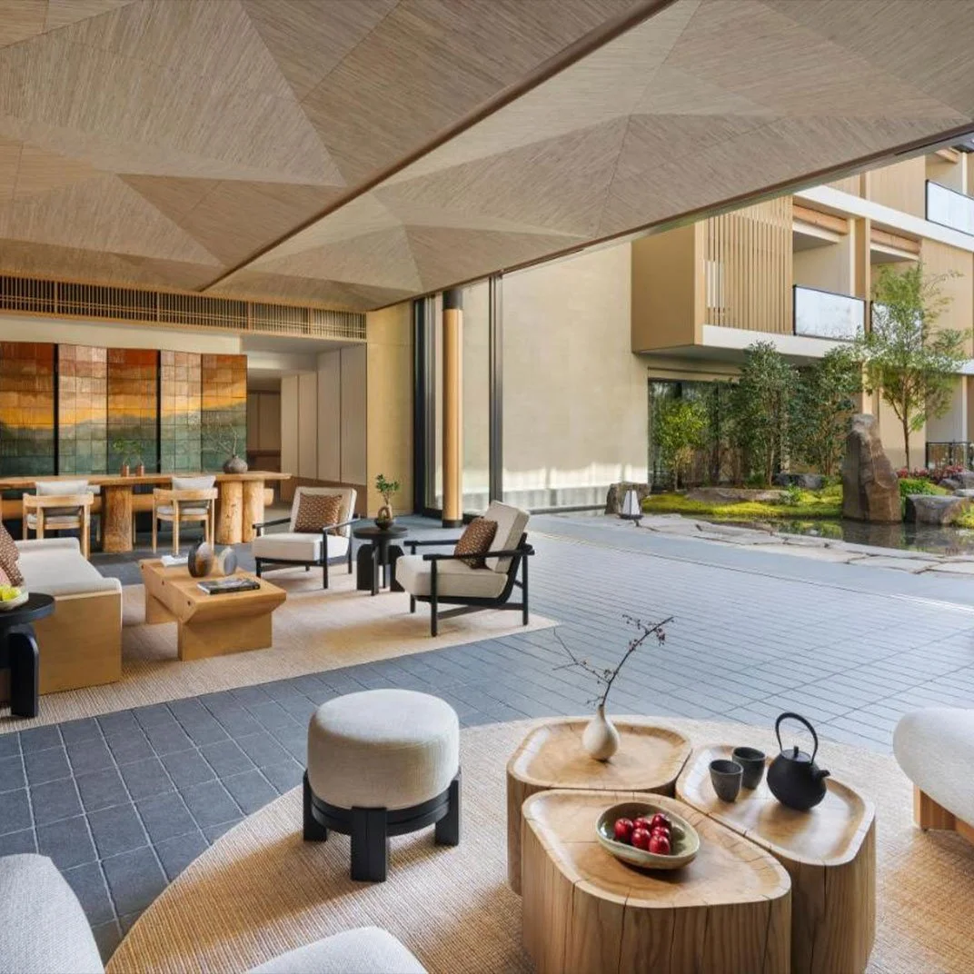 Modern hotel lobby with large windows, minimalist furniture, wooden decor, indoor plants, and a view of an outdoor garden.