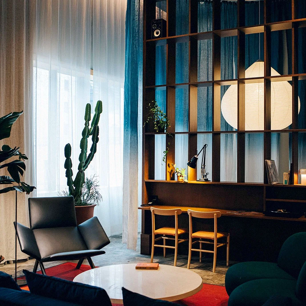 Living room with large window, sheer curtains, cactus plant, black modern armchair, round coffee table, bookshelf with decorative items, desk lamp, large paper lantern, decorative plants, and two wooden chairs.