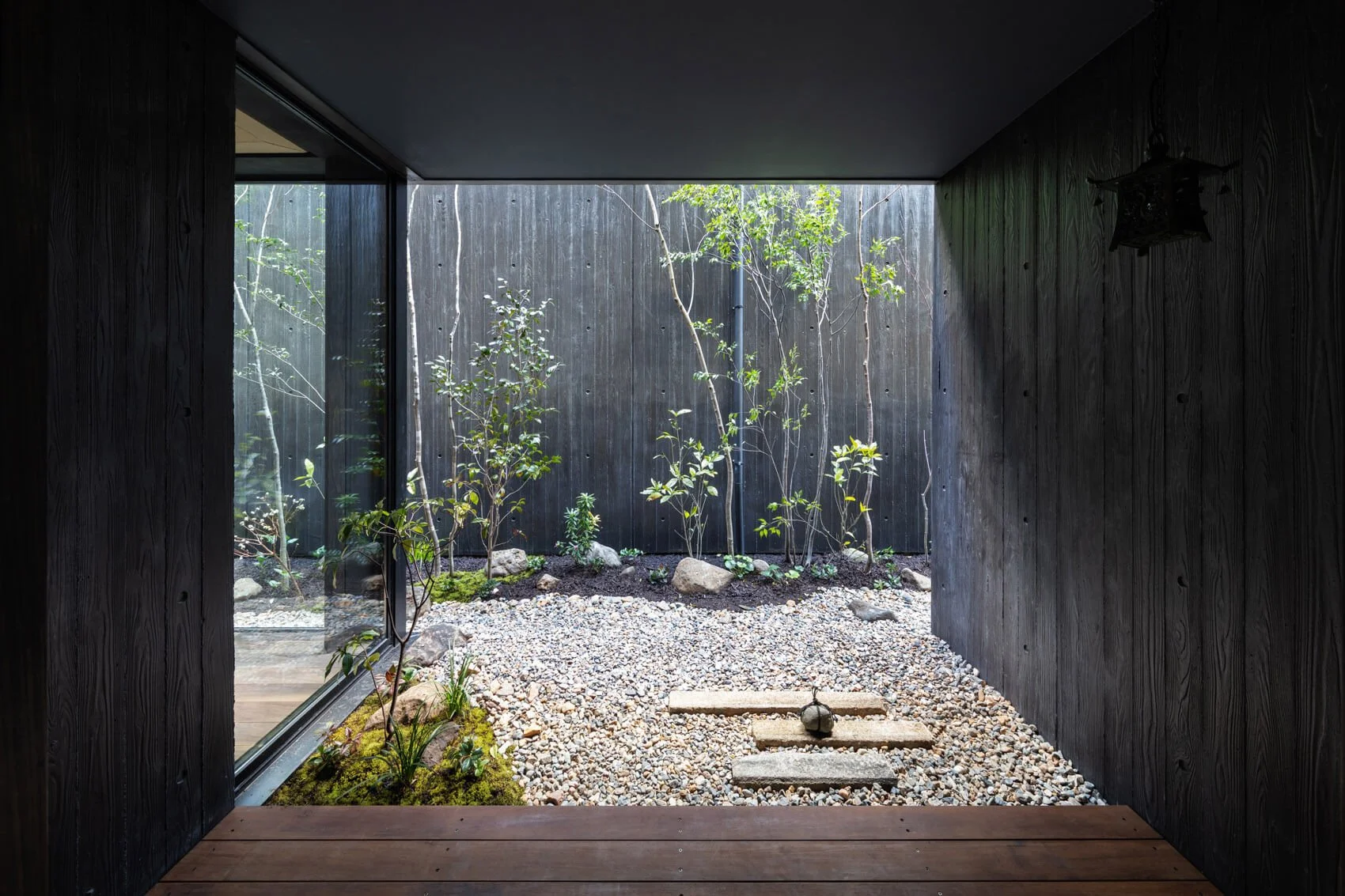 Interior view of a modern house looking out onto a small garden with rocks and small trees, with a dark wall on the right and a glass sliding door on the left.
