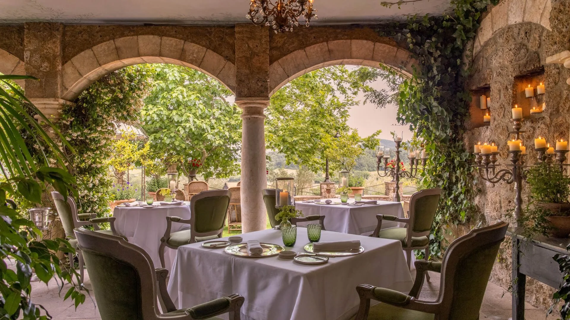 Elegant outdoor dining area with white tablecloths, green glassware, and candles, surrounded by greenery and stone arches.