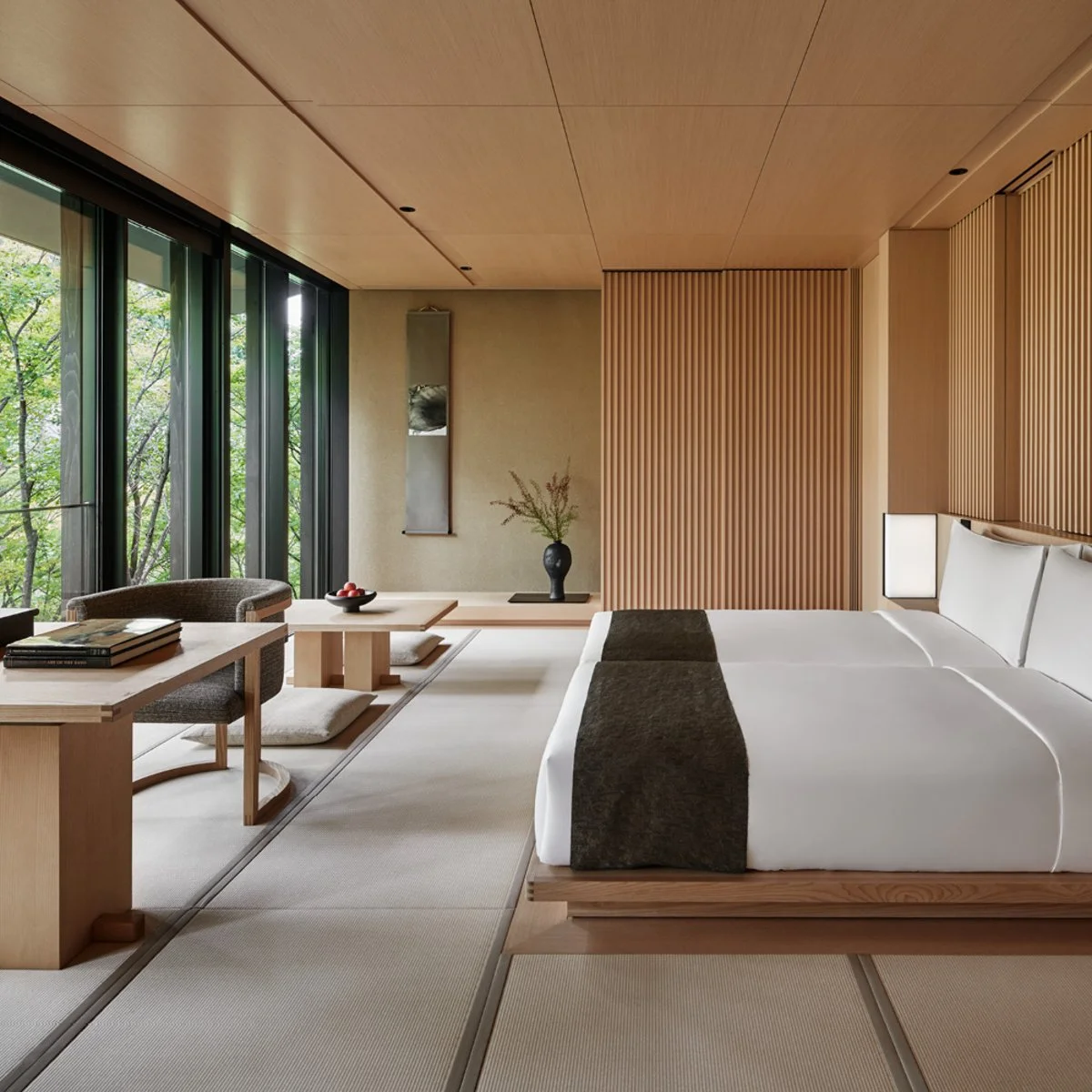 A modern Japanese-inspired hotel room with sliding glass doors, a bed with white linens, a dark throw, wooden wall paneling, a small table with books and cushions, a woven chair, and a vase with greenery, overlooking a green outdoor landscape.