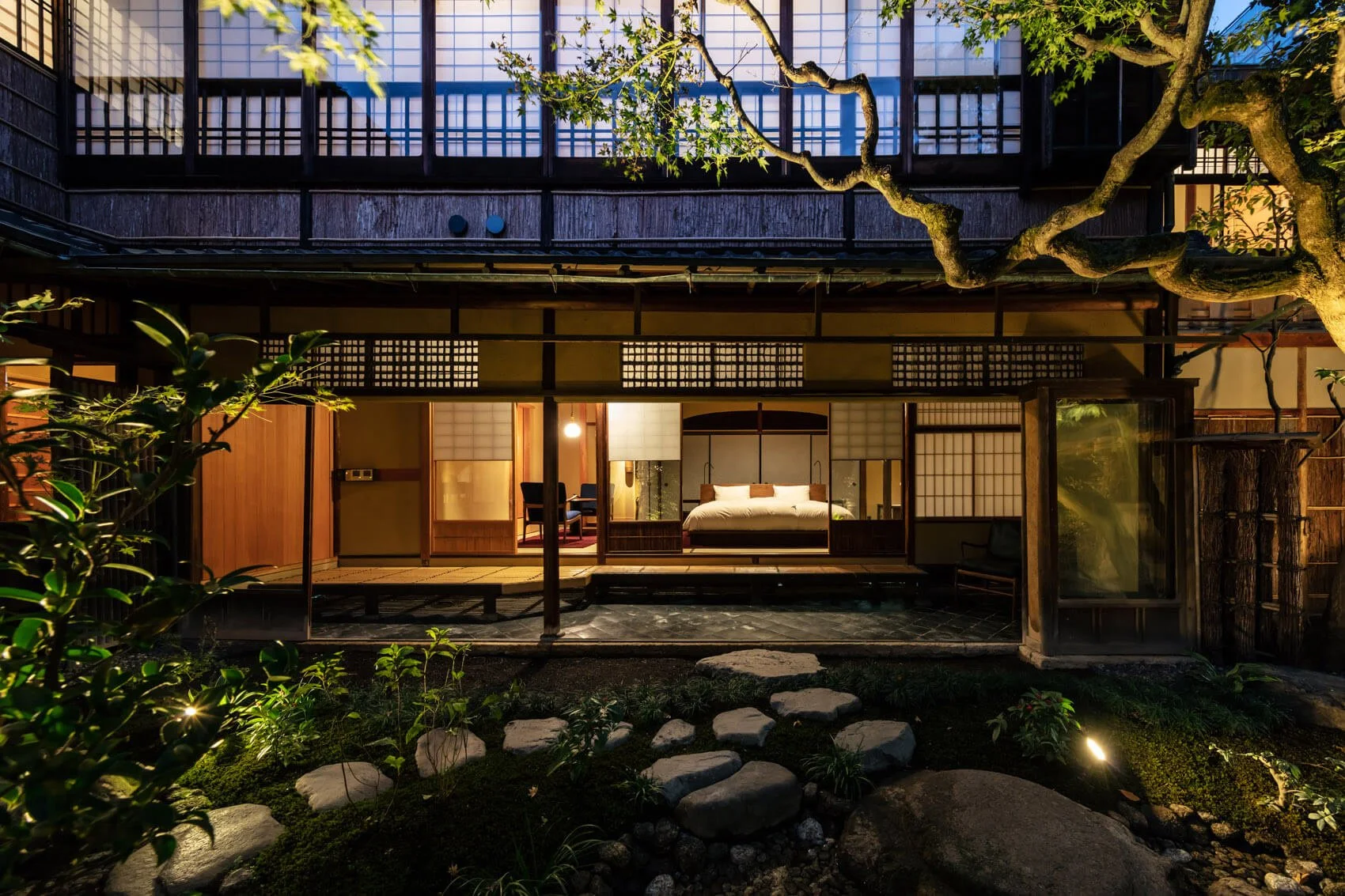 Exterior view of a traditional Japanese-style house at night, with an illuminated bedroom visible through sliding shoji doors, a garden with stepping stones and plants in the foreground, and a large tree arching over the house.