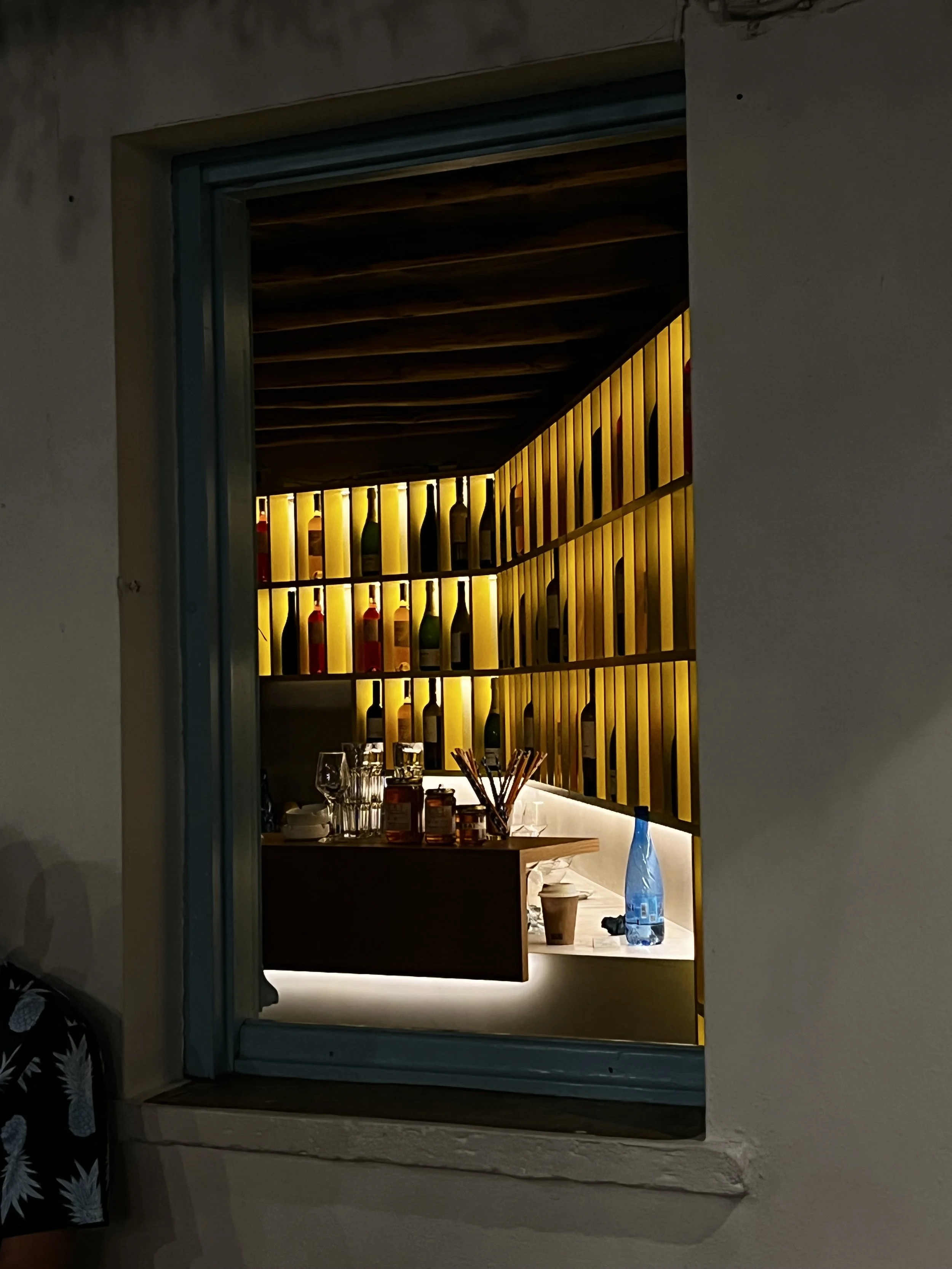 View through a window showing a wine bar with backlit shelves filled with wine bottles, glasses, and jars on a wooden counter.