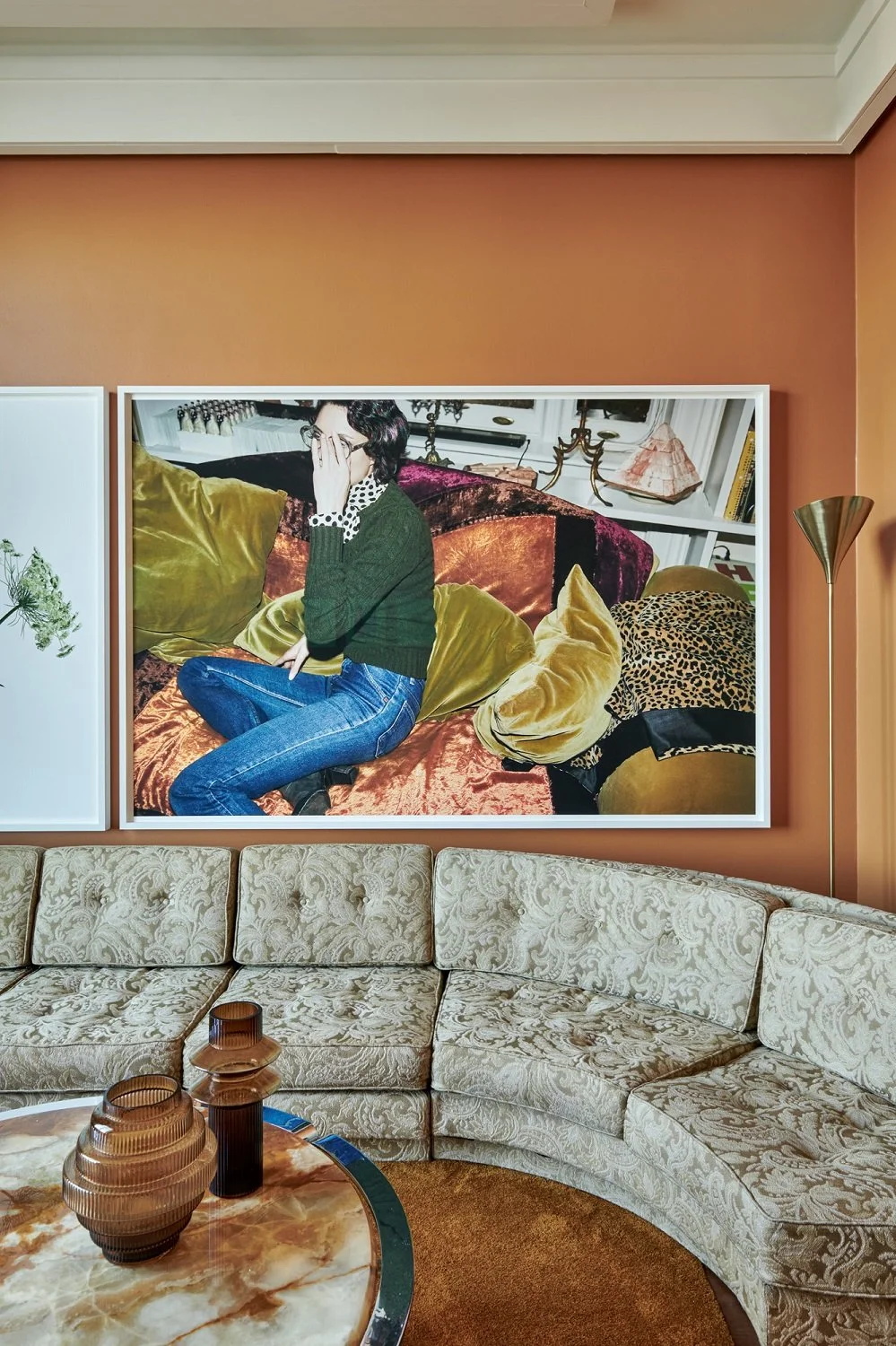 A framed photograph of a woman sitting on a luxurious velvet couch with gold, velvet, and leopard print pillows. The woman has dark hair, wears glasses, a dark green sweater with a polka dot shirt, and blue jeans, and is covering her mouth with her hand. The background features a white bookshelf and decorative items.