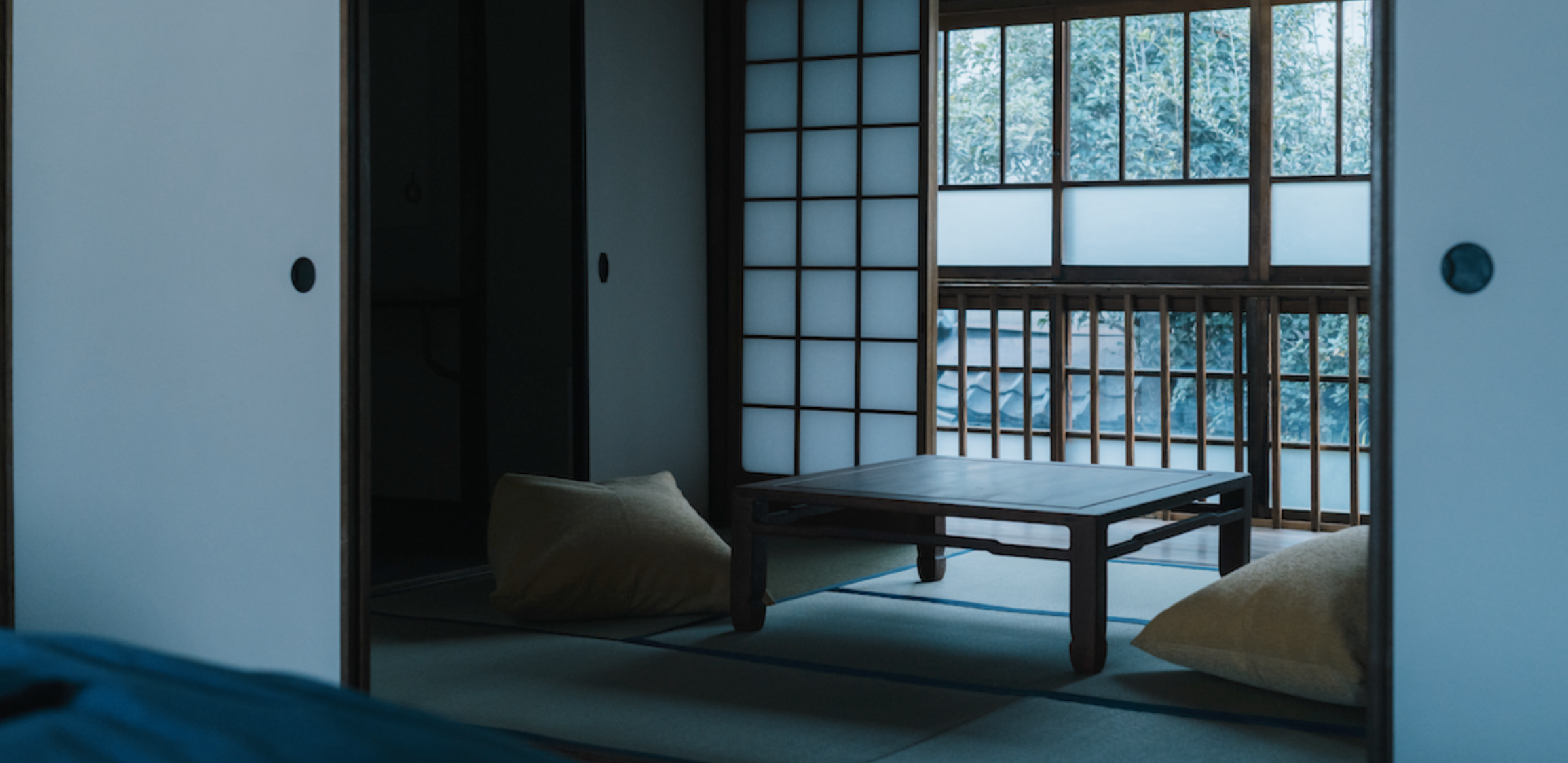 A traditional Japanese tatami room with shoji sliding doors and wooden balcony overlooking greenery. The room has floor cushions and a low wooden table.