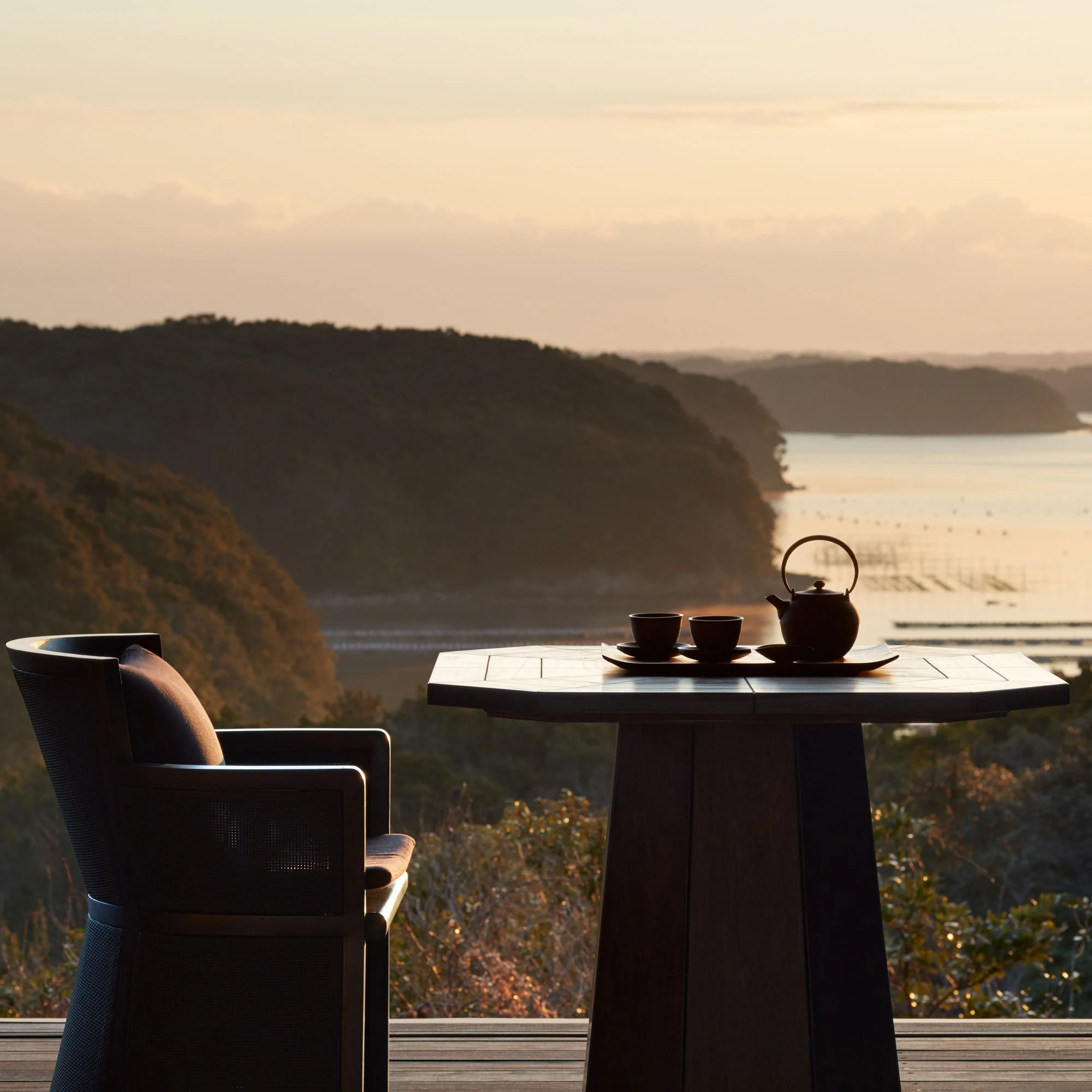 A cozy outdoor seating area with a wooden table, a teapot, two cups, and a tray, overlooking a serene bay with hills during sunset.