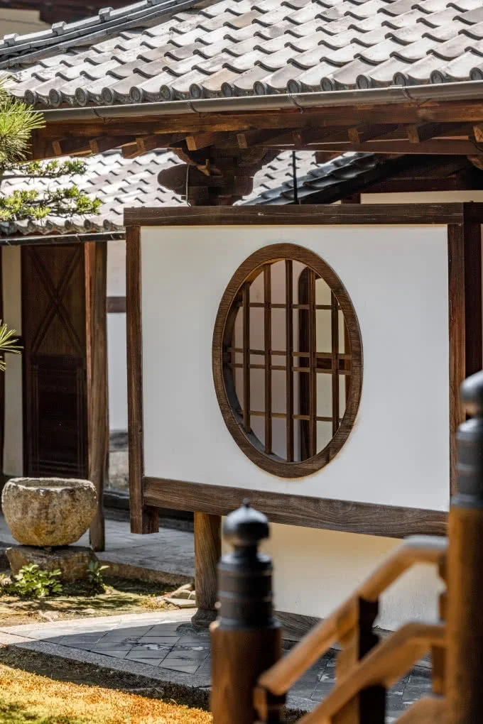 A traditional Japanese-style building with a tiled roof, white walls, and a circular wooden window with grid pattern, surrounded by garden elements.