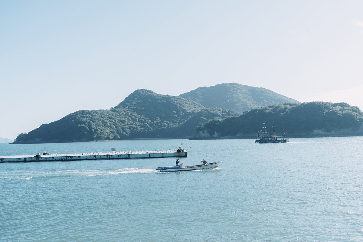 A serene sea scene with mountains in the background, a boat moving across the water, and a ship near the horizon.