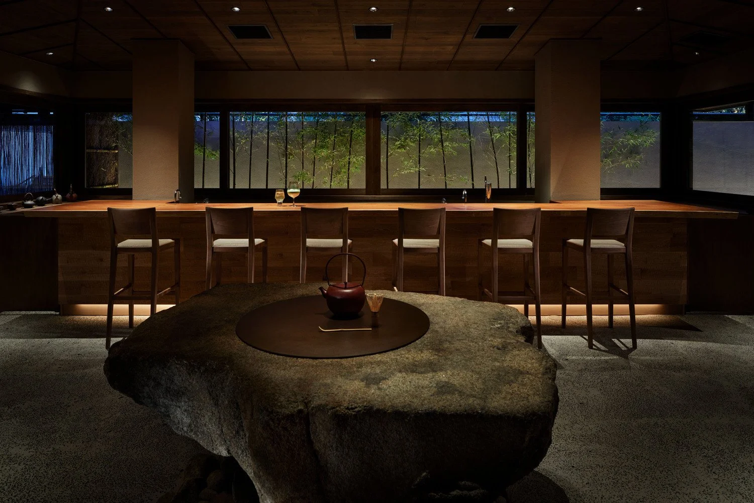 Interior of a modern, dimly lit dining area with a long wooden bar, six bar stools, a large stone table with a teapot and a glass on top, and large windows showing greenery outside.