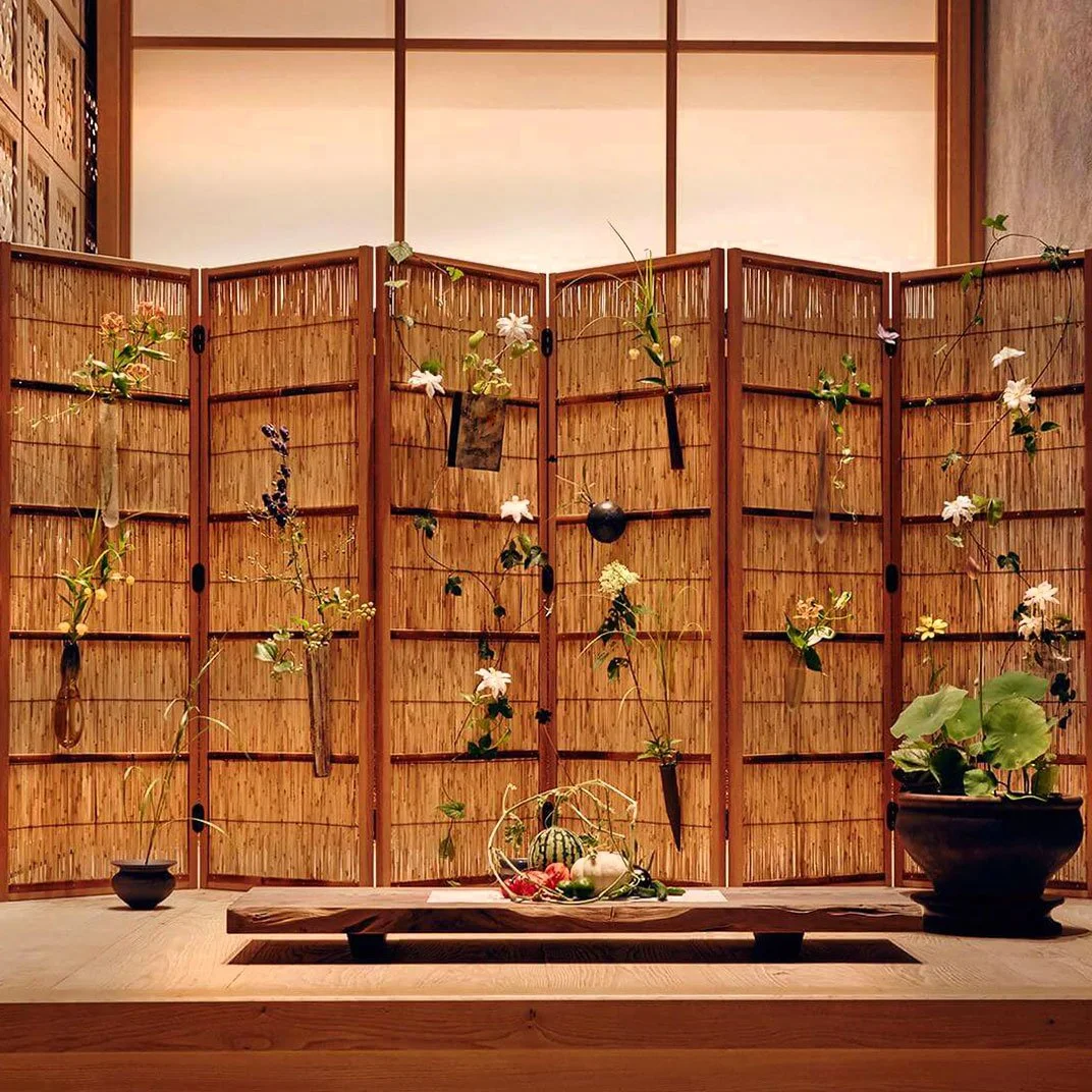 Decorative Japanese-style room with a bamboo folding screen, hanging flowers, and a low wooden table with fruit and vegetables, and a pot with green leafy plants.
