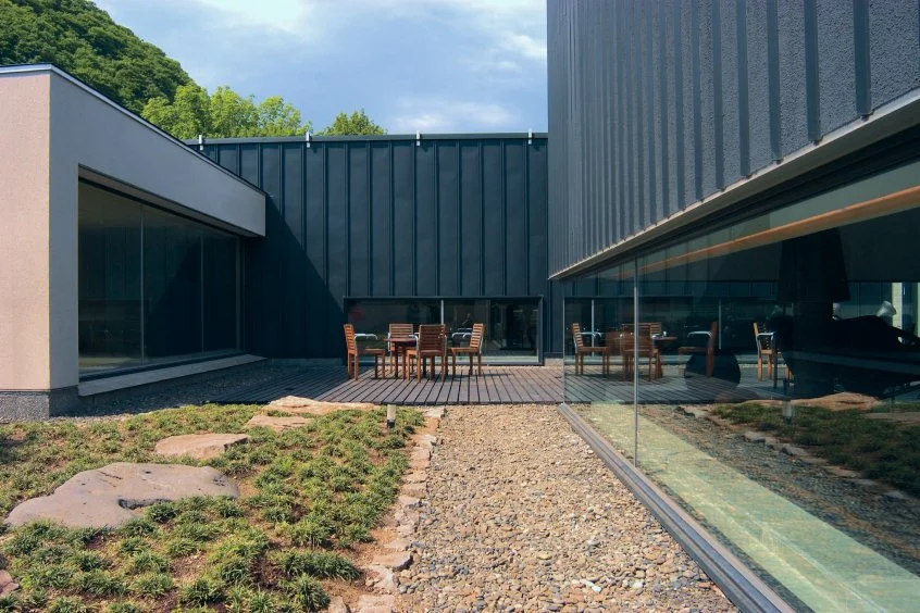 Modern building with outdoor patio, wooden table and chairs, large glass windows reflecting the patio, gravel and patchy grass ground, and green trees in the background.