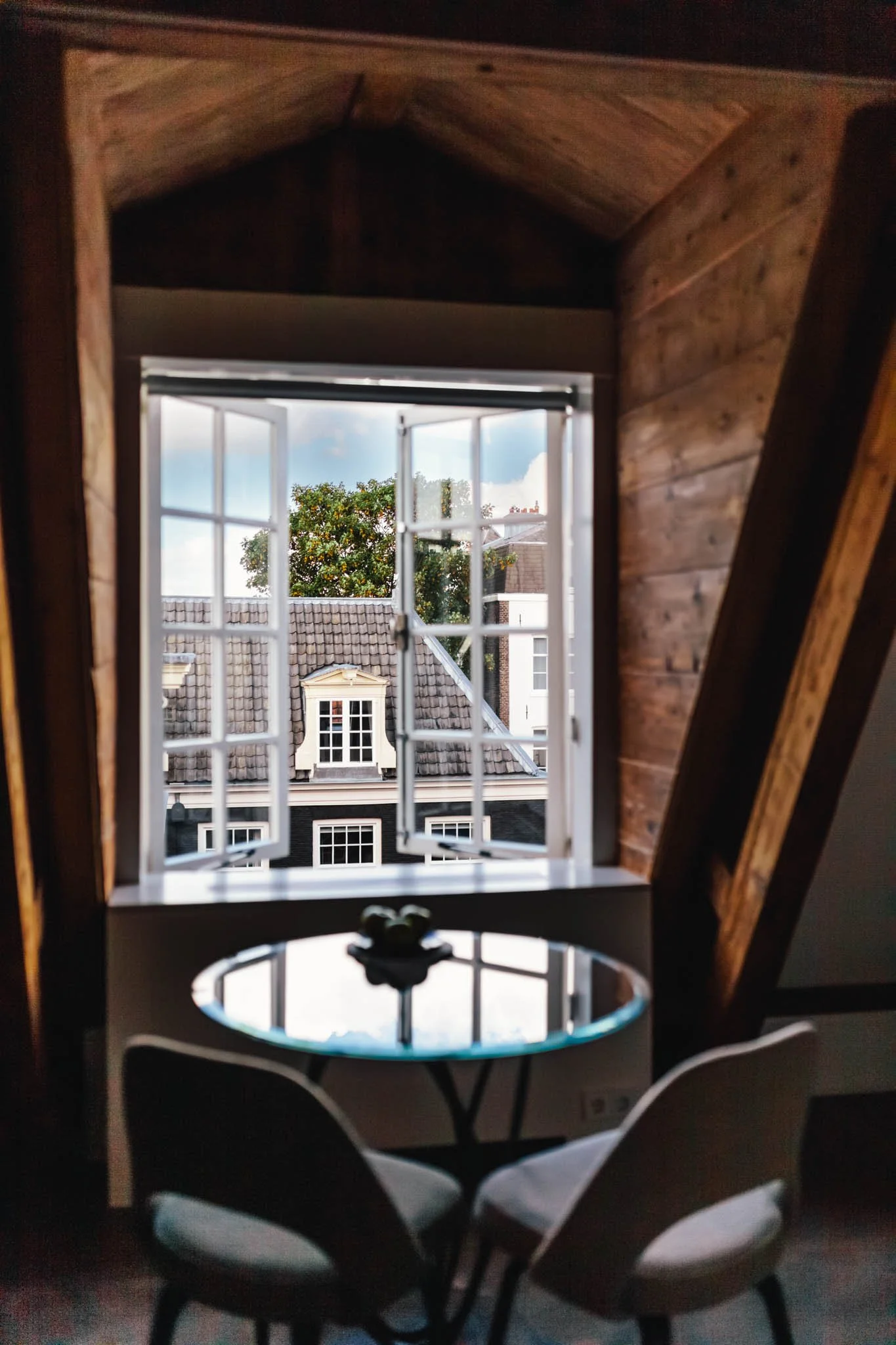 An interior view of a cozy attic room with wooden walls and ceiling, a small round glass table with chairs, and an open window showcasing rooftops and a tree outside.
