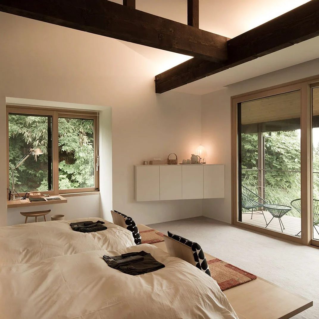 A bedroom with two beds, a window with green trees outside, a sliding glass door leading to a balcony with outdoor chairs, a white wall-mounted cabinet, and wooden beams on the ceiling.