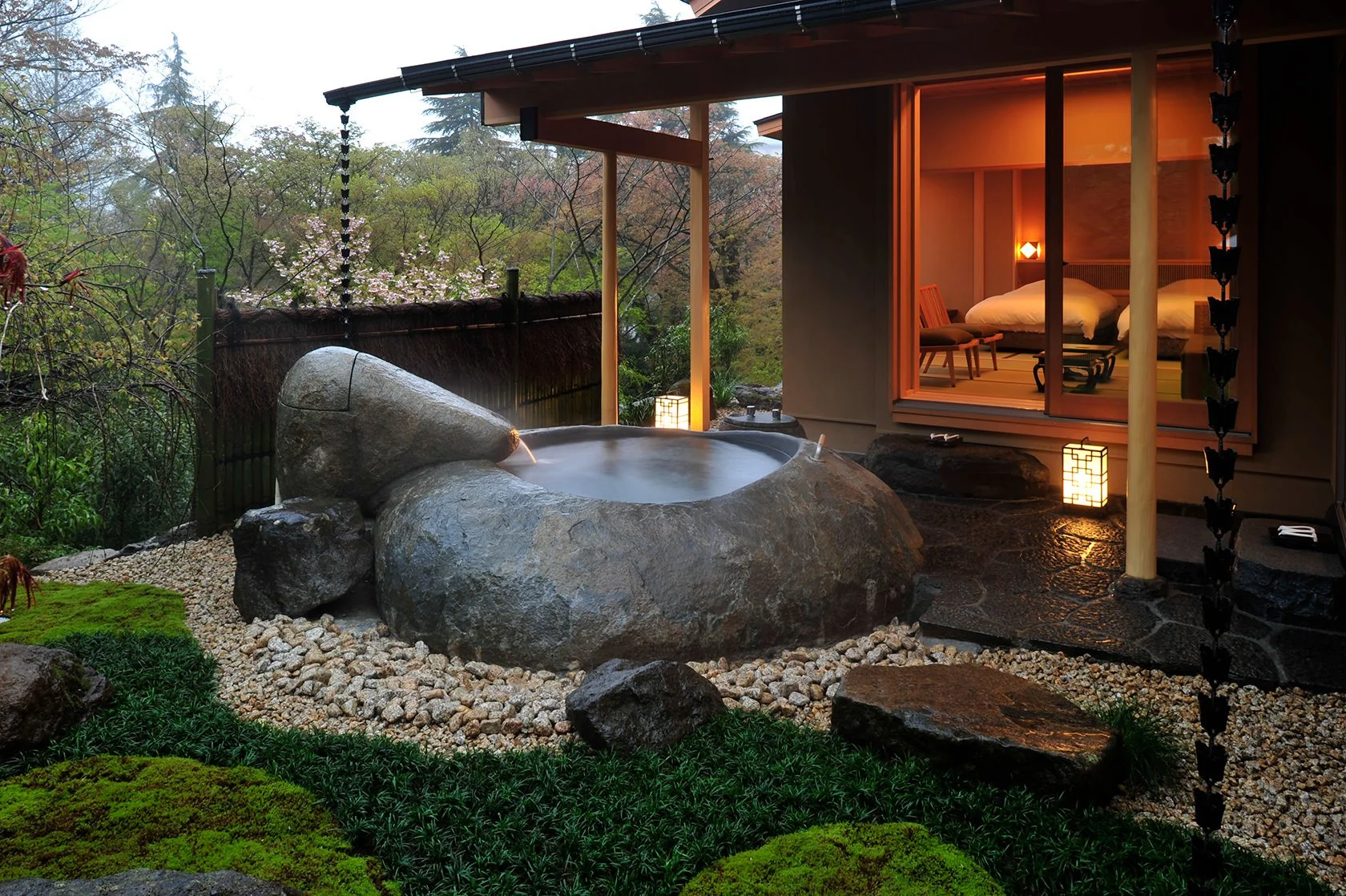A traditional Japanese-style outdoor onsen bath with large rocks, surrounded by a lush garden with moss and greenery, adjacent to a modern room with sliding glass doors, warm interior lighting, and seating inside.