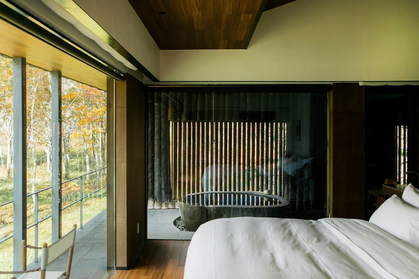A modern bedroom with a large window showing trees outside, a sliding glass door leading to a balcony, and a black bathtub on the balcony with wooden slats providing privacy.