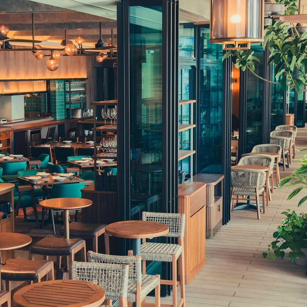 A modern restaurant interior with round wooden tables, cushioned chairs, and glass partitions. The space is decorated with warm lighting, plants, and teal-colored seating.