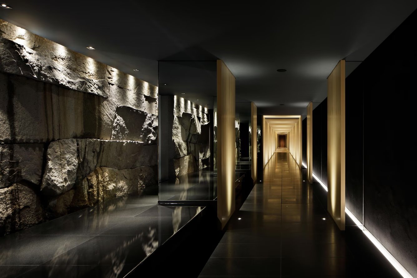 Modern hotel corridor with stone wall on the left, black and gold accents, and warm lighting, leading to a distant door.