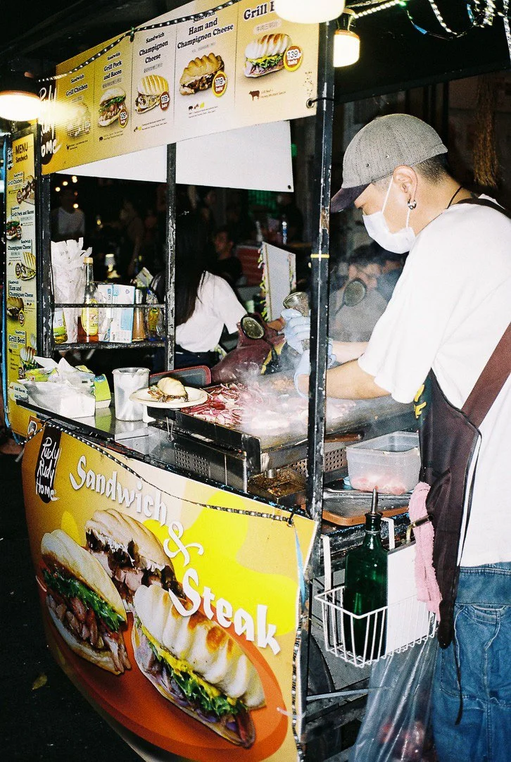 A street food stall that serves sandwiches and steak, with a menu board displaying various sandwich options. A chef wearing a face mask, gloves, and a gray cap is grilling meat on a stove.