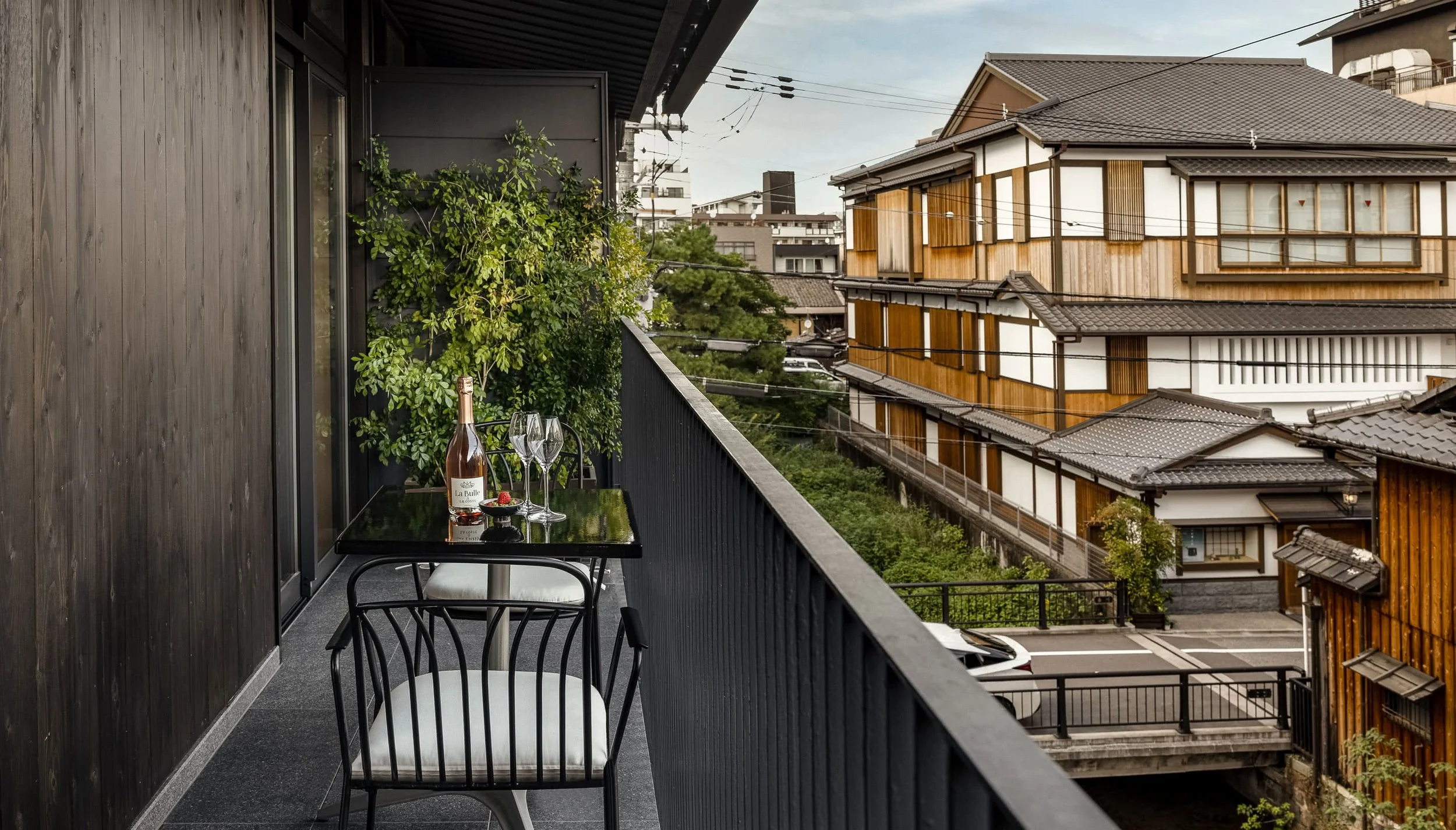 A balcony with a black table set with a bottle of rosé wine and two glasses, overlooking a street with traditional Japanese wooden buildings and greenery.