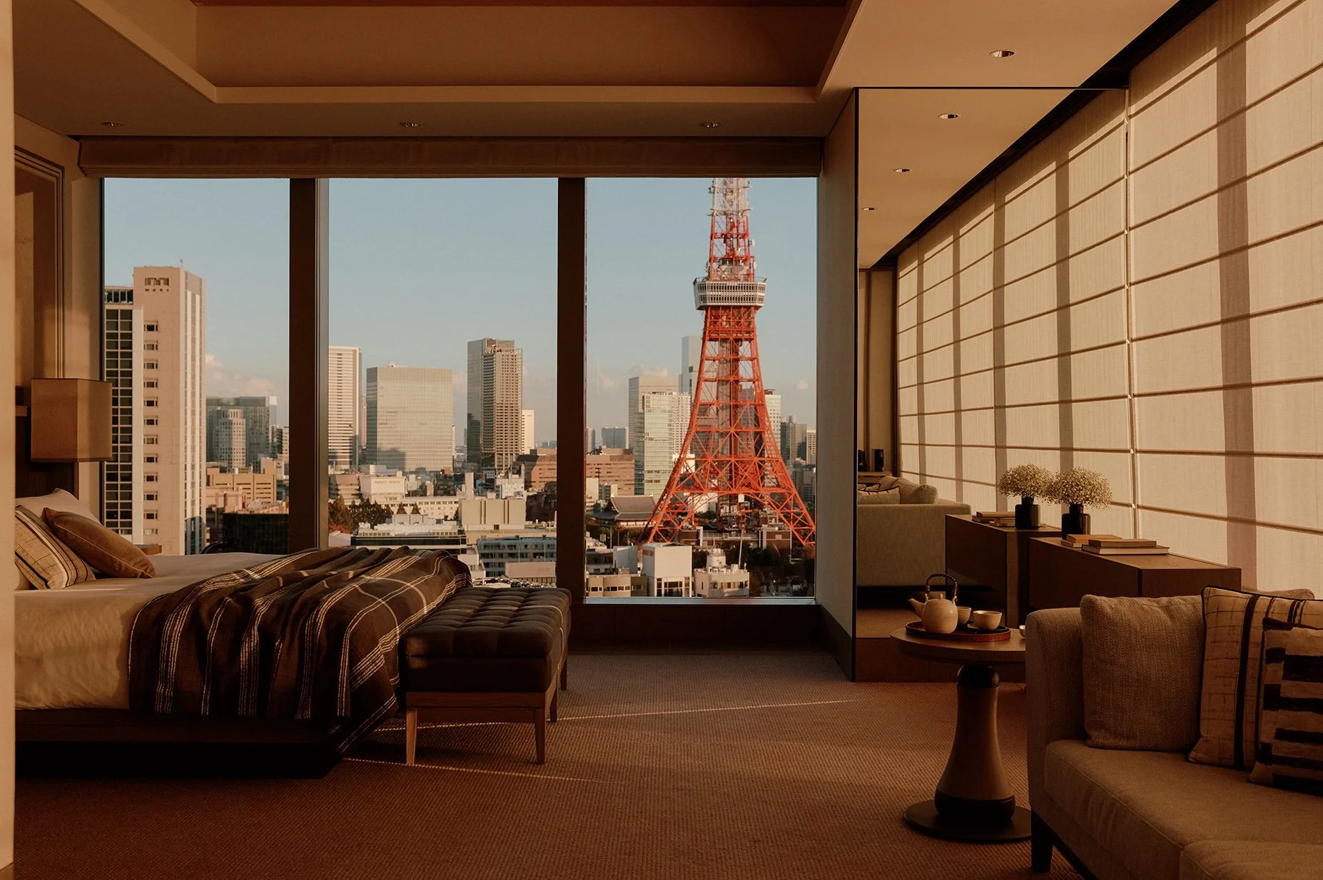 Luxury hotel room with large windows showing Tokyo Tower and city skyline during the day, with warm lighting and modern decor.