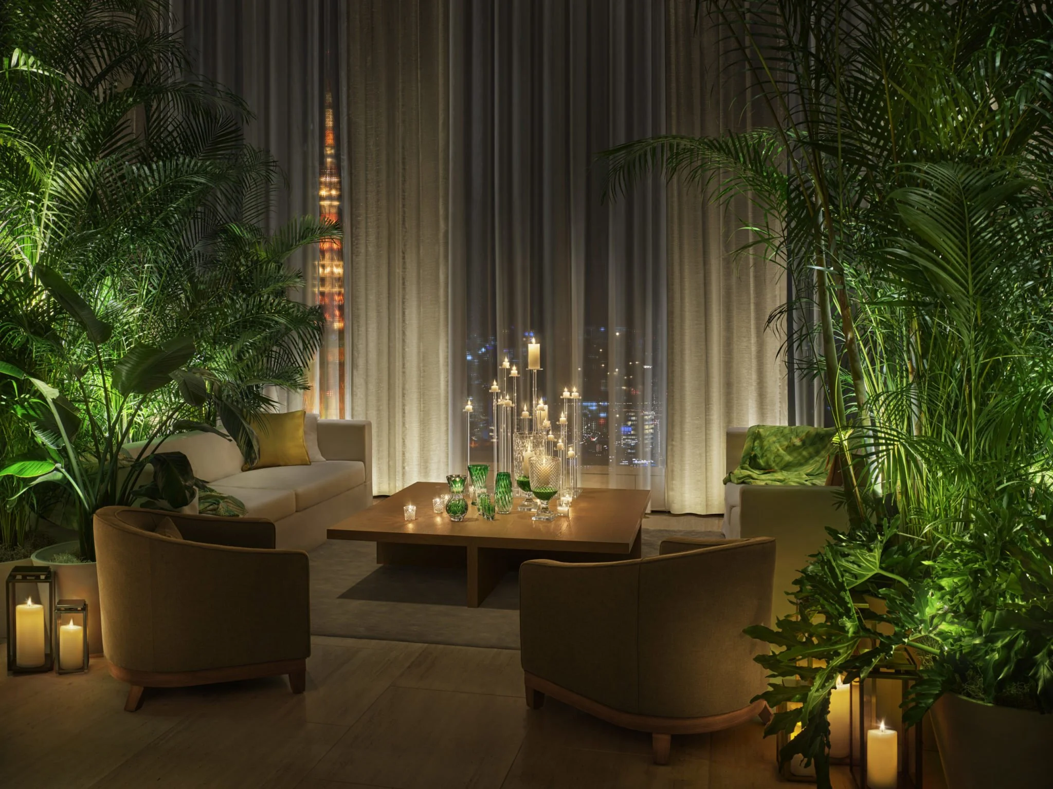 A cozy living room with lush green plants, candles, a wooden table, and large windows with curtains at night.