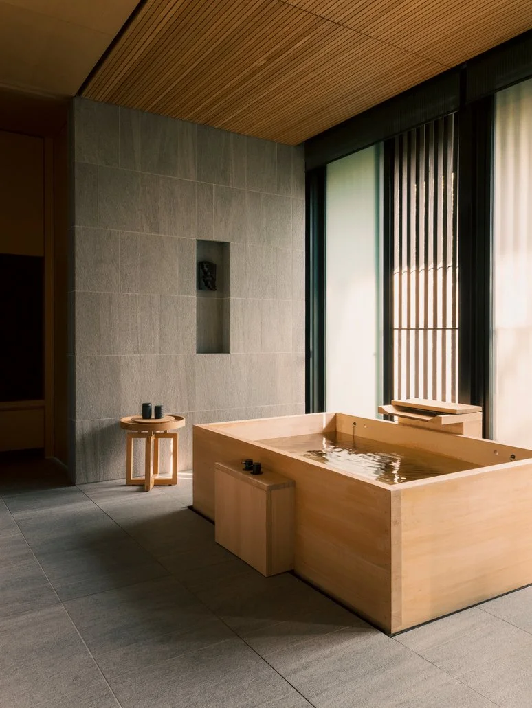 A minimalist indoor space with a wooden soaking tub filled with water, a small wooden stool with two black cups, and a large window with vertical blinds allowing natural light to enter. The room has gray tiled flooring, a gray tiled wall with a recessed niche, and a wooden ceiling.