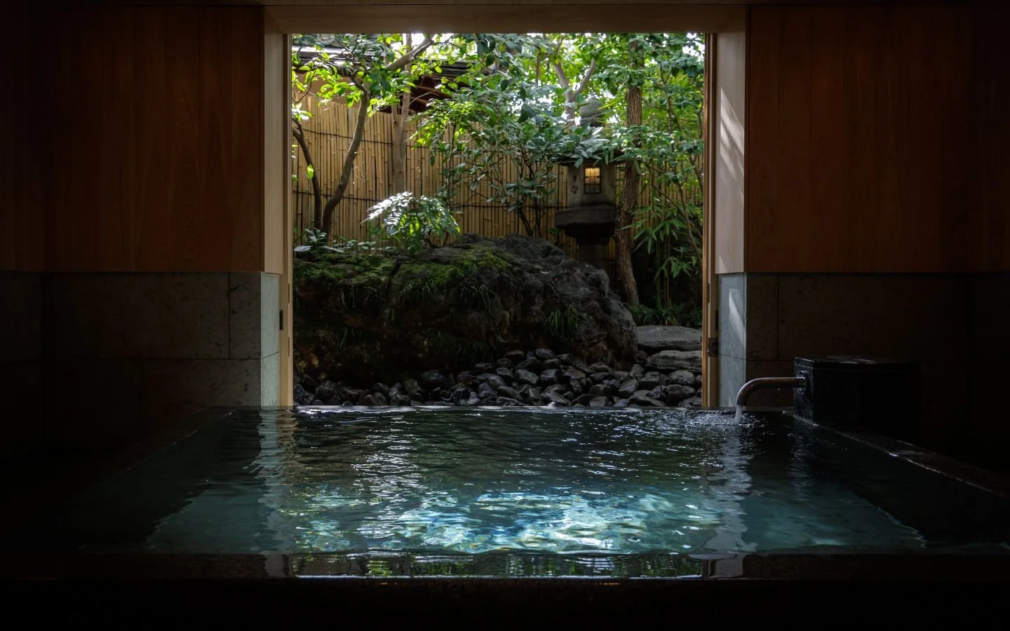A traditional Japanese-style indoor bath with a view of a serene outdoor garden featuring rocks, trees, and a stone lantern.
