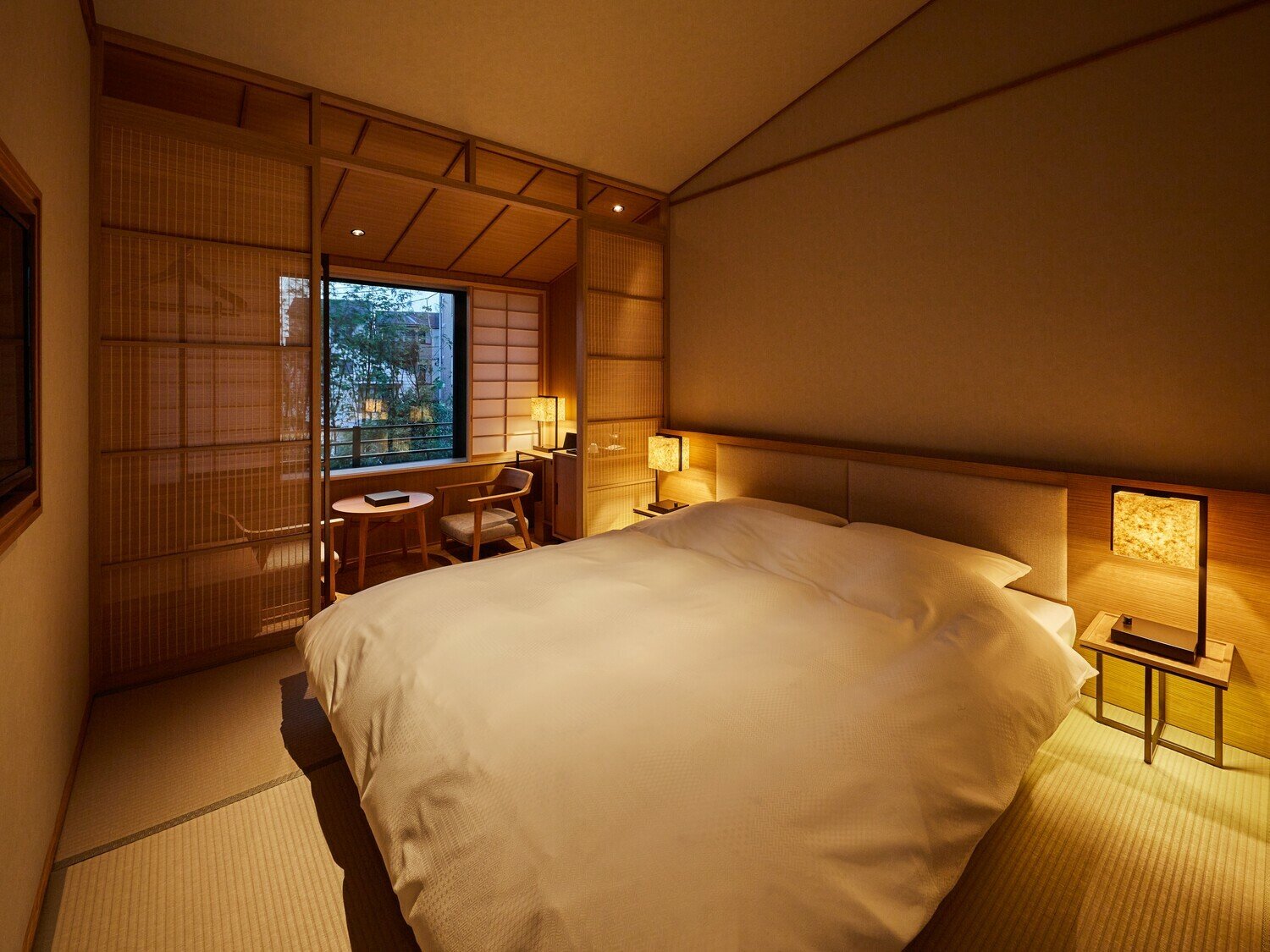 A cozy hotel room with a large bed, side tables with lamps, a window overlooking greenery, and traditional Japanese shoji screens.