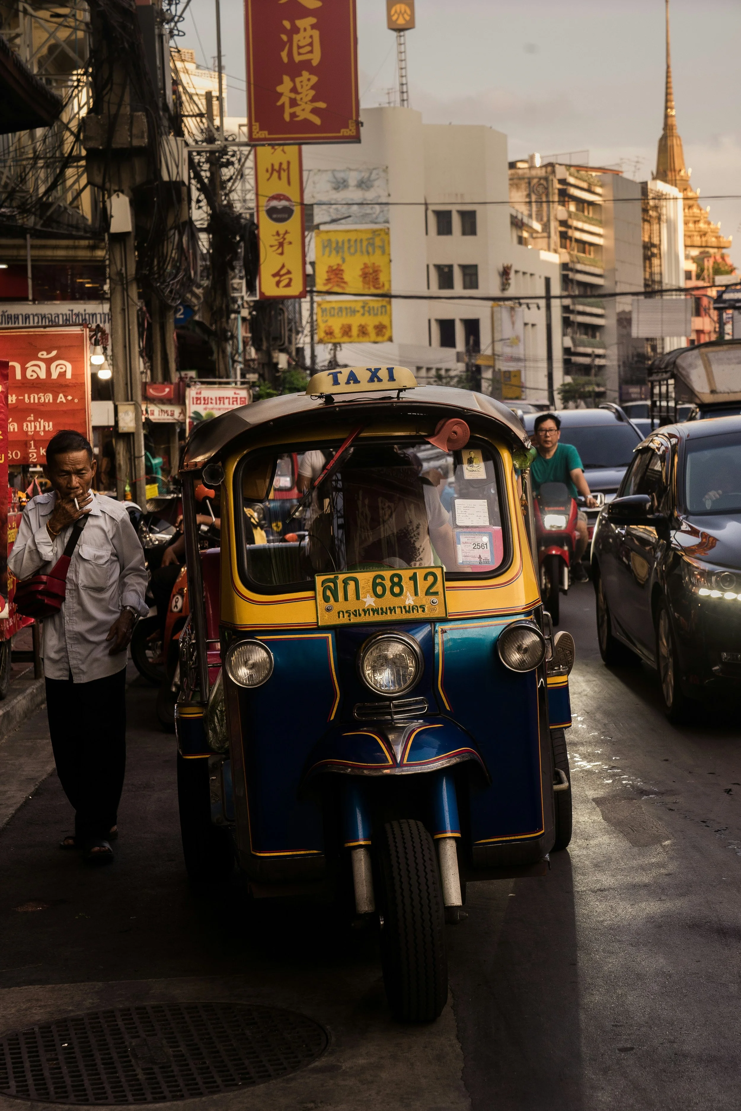 A colorful tuk-tuk parked on a busy street in Bangkok with buildings and signage in the background.