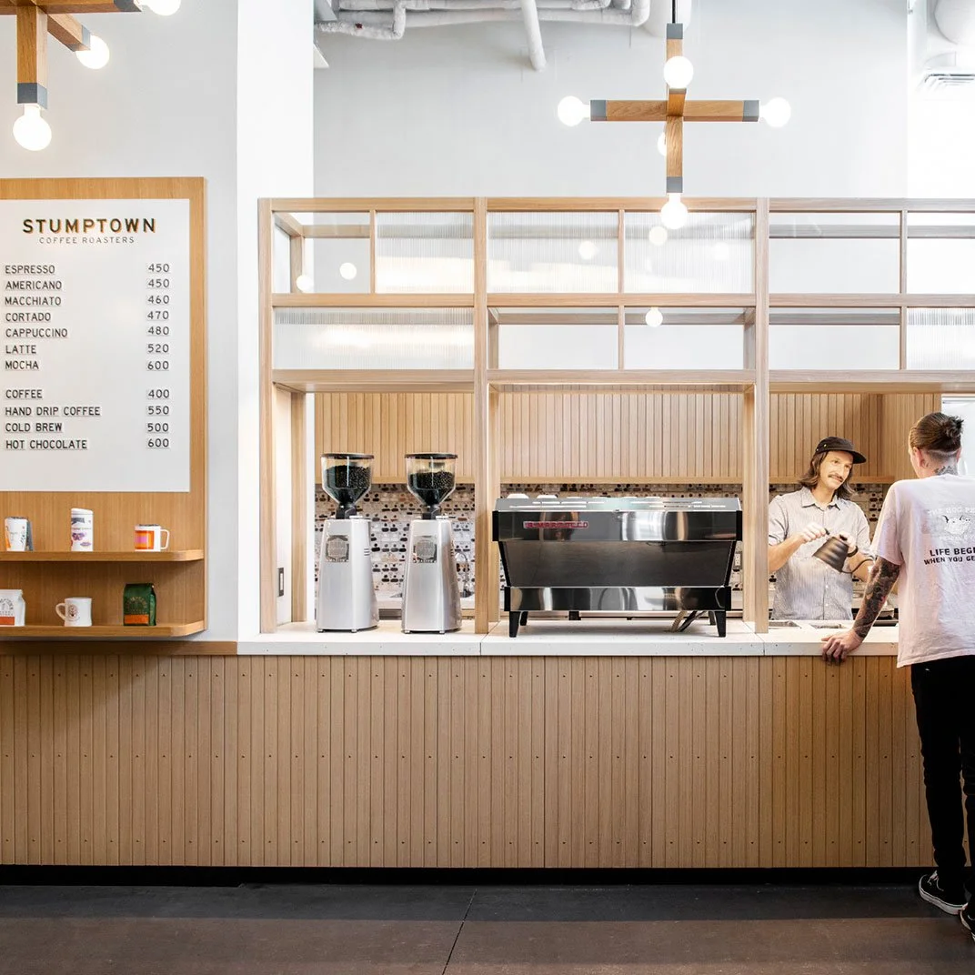 Interior of a modern coffee shop with a wooden counter, coffee menu on a white board, two coffee grinders, an espresso machine, and a barista talking to a customer.