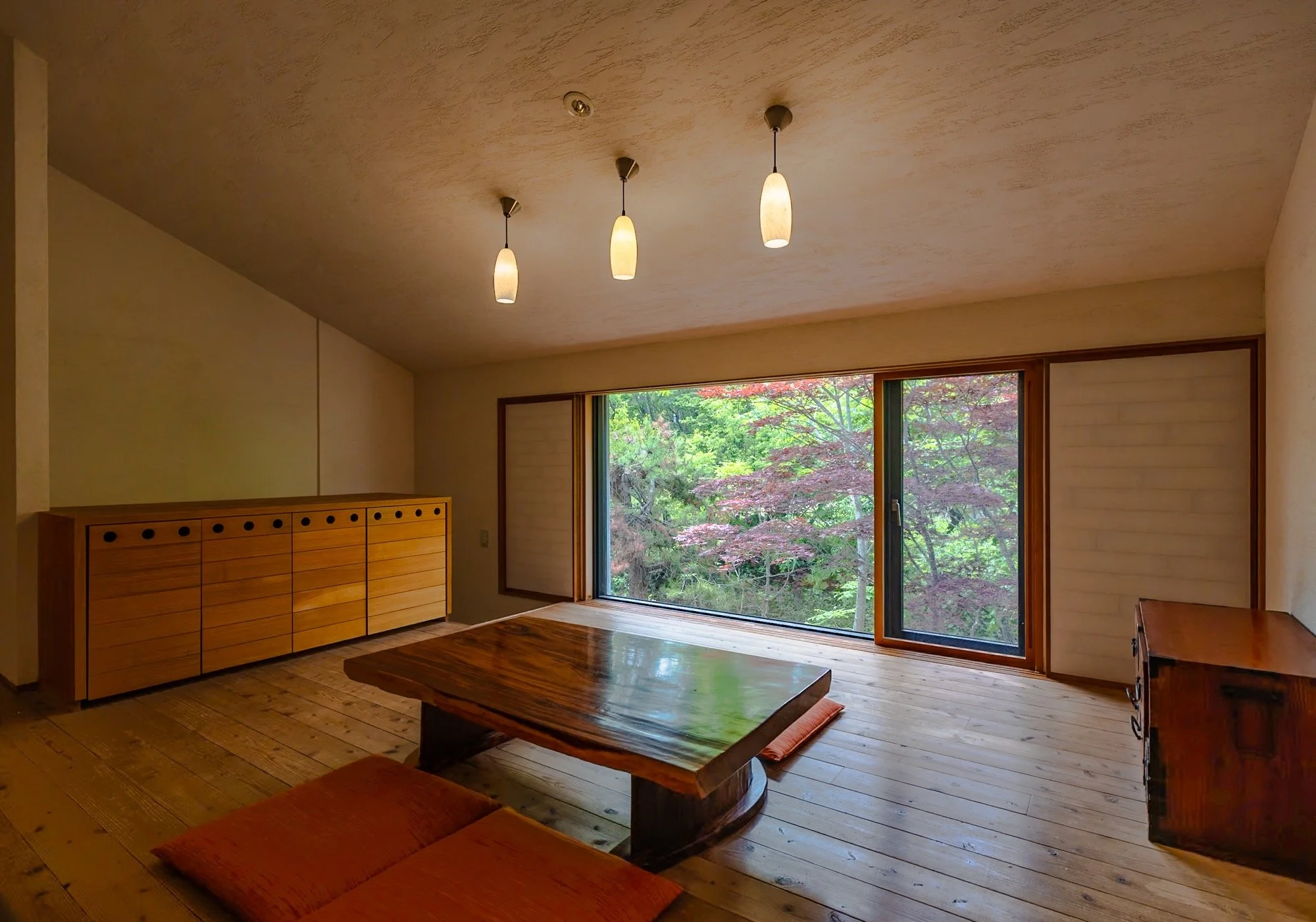 Interior of a minimalistic room with wooden flooring, a low wooden table, two cushions, a wooden cabinet, and large sliding glass doors opening to a view of green trees and pink flowers.