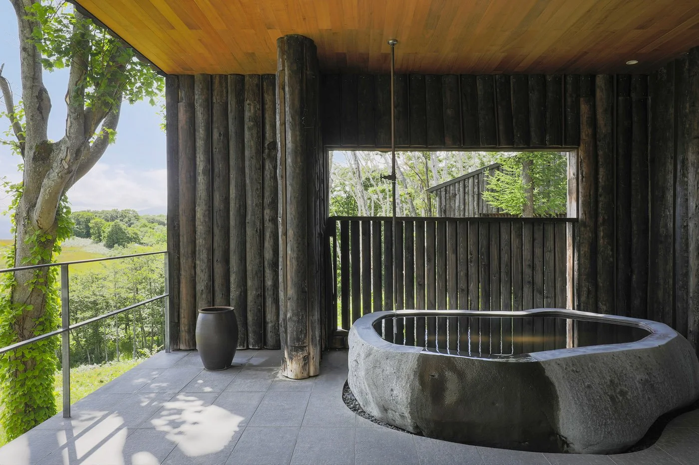A rustic outdoor bathing area featuring a smooth stone bathtub filled with water on a tiled floor, surrounded by weathered dark wood walls and a ceiling, with a large open window revealing lush green trees and landscape.