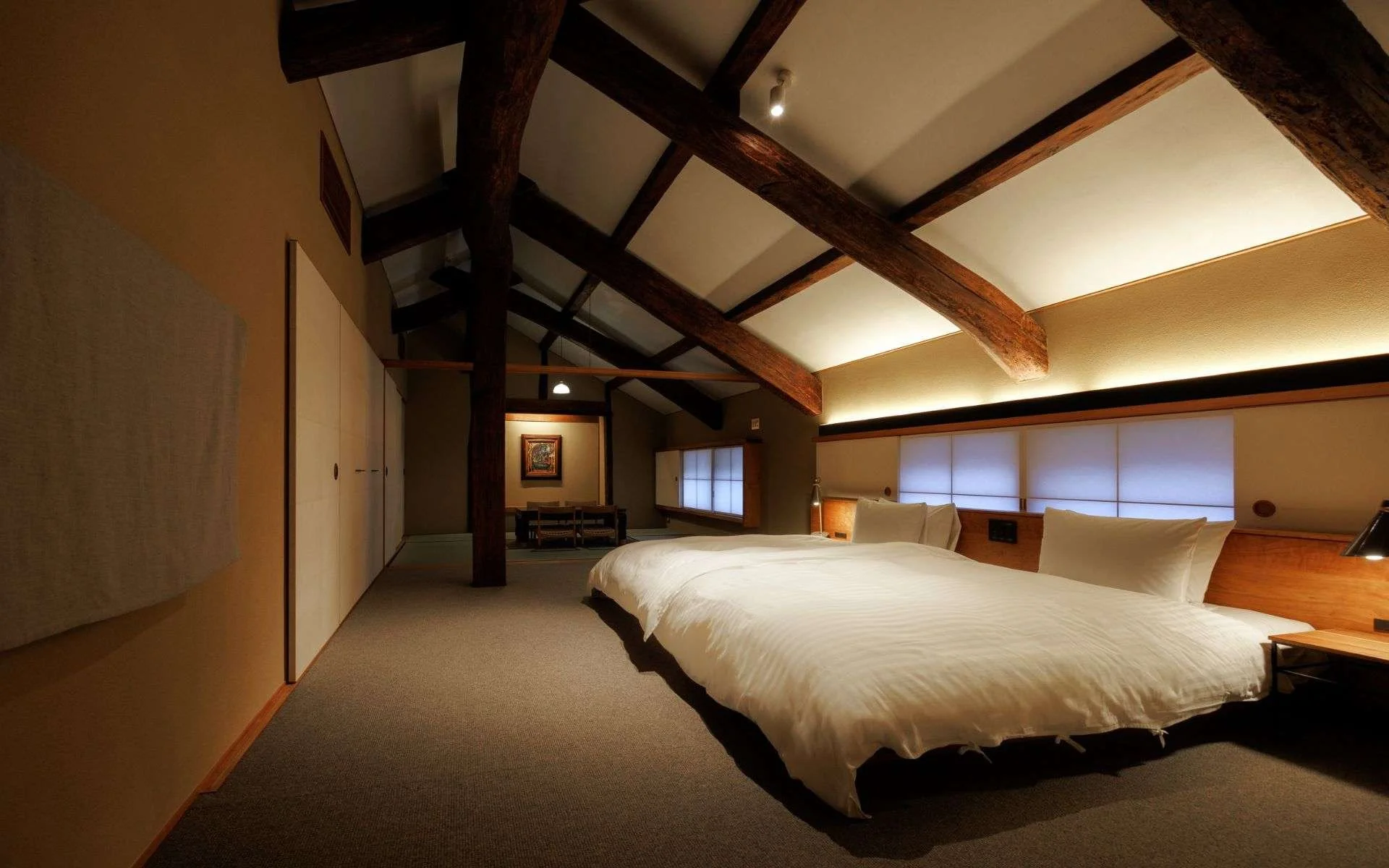 A cozy hotel bedroom with a large bed, wooden beams on the ceiling, and warm lighting.
